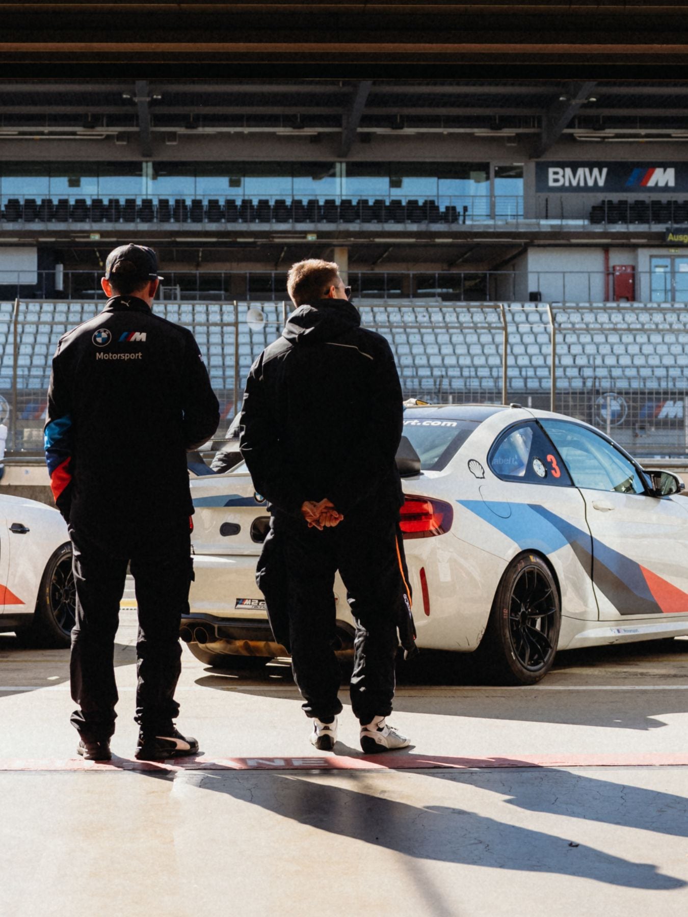 Zwei Personen in schwarzen BMW Motorsport-Jacken stehen auf einer Rennstrecke und betrachten einen weißen BMW-Rennwagen mit Rennlackierung. Im Hintergrund sind leere Tribünen und BMW-Branding an den Streckenwänden zu sehen.