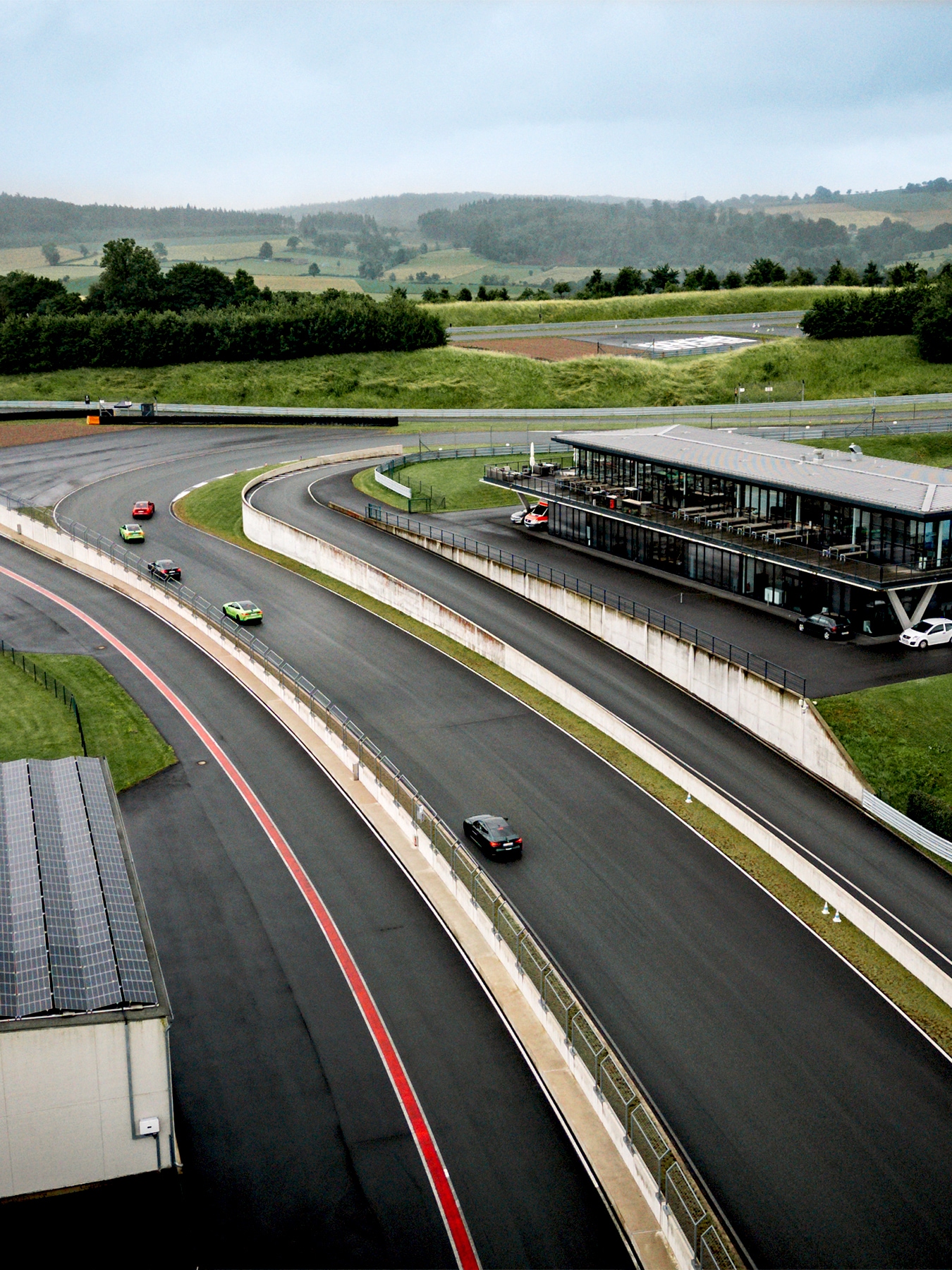 Eine Luftaufnahme einer Motorsport-Rennstrecke mit mehreren bunten Autos auf der Fahrbahn. Auf der rechten Seite ist ein modernes Boxengebäude mit Zuschauerbereich zu sehen. Die Rennstrecke ist von üppiger grüner Landschaft umgeben, mit sanften Hügeln und Wäldern im Hintergrund unter einem bewölkten Himmel.