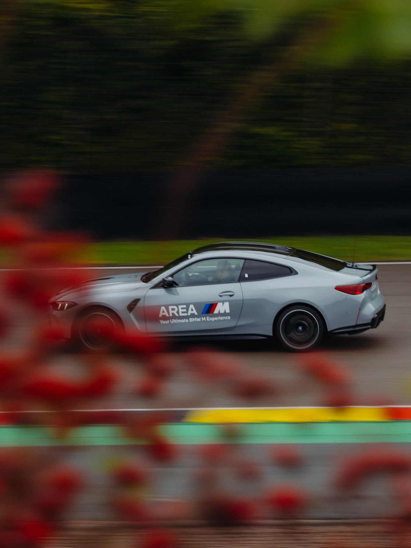 Ein silbernes BMW M Serienfahrzeug in Bewegung auf einer Rennstrecke. Das Fahrzeug trägt die Aufschrift 'AREA M' an der Seite mit den charakteristischen BMW M-farbigen Streifen. Der Hintergrund ist verschwommen mit roten, grünen und gelben Streifen, was auf eine Hochgeschwindigkeitsbewegung hindeutet. Das Fahrzeug hat ein elegantes Coupé-Design mit einem Heckspoiler.