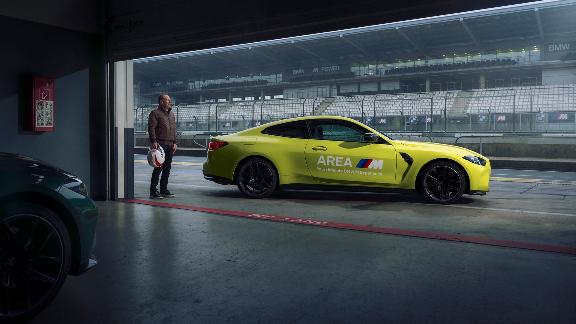Ein leuchtend gelber BMW M Sportwagen mit 'AREA'-Branding, der in einer Renngarage parkt, wobei eine Rennstrecke durch die offene Tür sichtbar ist. Eine Person in brauner Jacke steht neben dem Auto und hält einen Helm. Das BMW M Power Branding ist auf den Barrieren entlang der Strecke zu sehen.