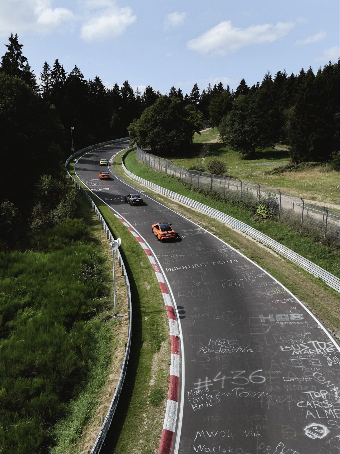 Luftaufnahme der Nürburgring-Rennstrecke mit mehreren Sportwagen, die entlang einer Kurve fahren. Die Strecke ist von dichtem Wald umgeben und hat rot-weiße Randsteine. Verschiedene Botschaften und Graffiti sind auf der Asphaltoberfläche sichtbar, darunter deutlich geschrieben 'NÜRBURG TEAM'.