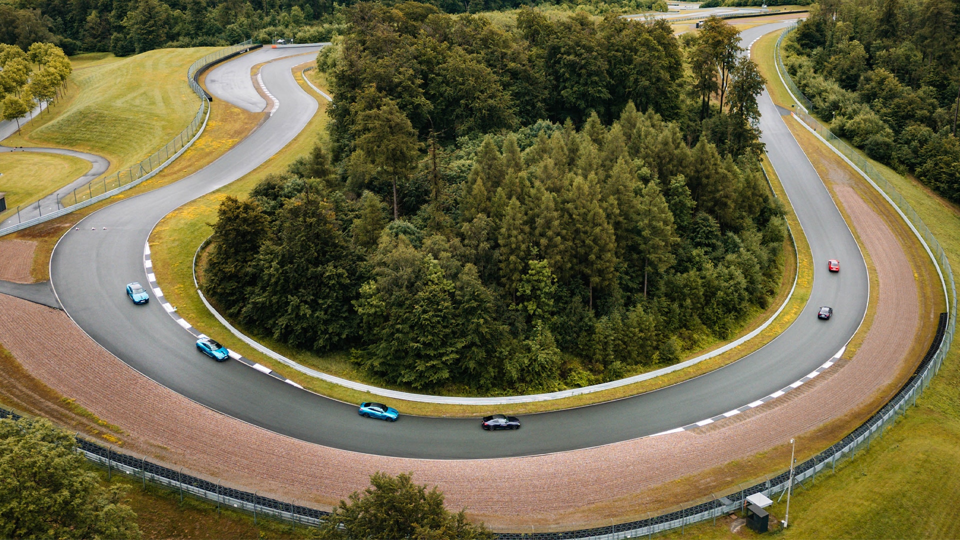 Luftaufnahme einer kurvenreichen Rennstrecke oder Teststrecke mit mehreren bunten Fahrzeuge, die die Kurven befahren. Die Strecke weist scharfe Kurven um ein zentrales Waldgebiet auf, mit grauer Asphaltoberfläche, die von Kiesflächen und grünem Gras umgeben ist. Die Rennstrecke ist von Wald umgeben.