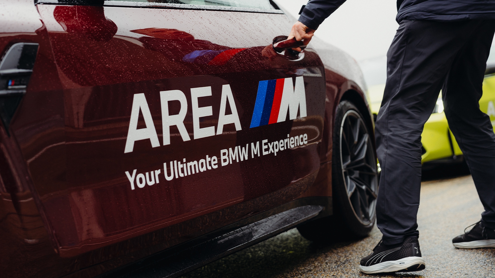 Ein rotes BMW-Fahrzeug mit der Aufschrift 'AREA M' und 'Your Ultimate BMW M Experience' auf der Seitentür. Eine Person in dunkler Kleidung öffnet die Autotür.