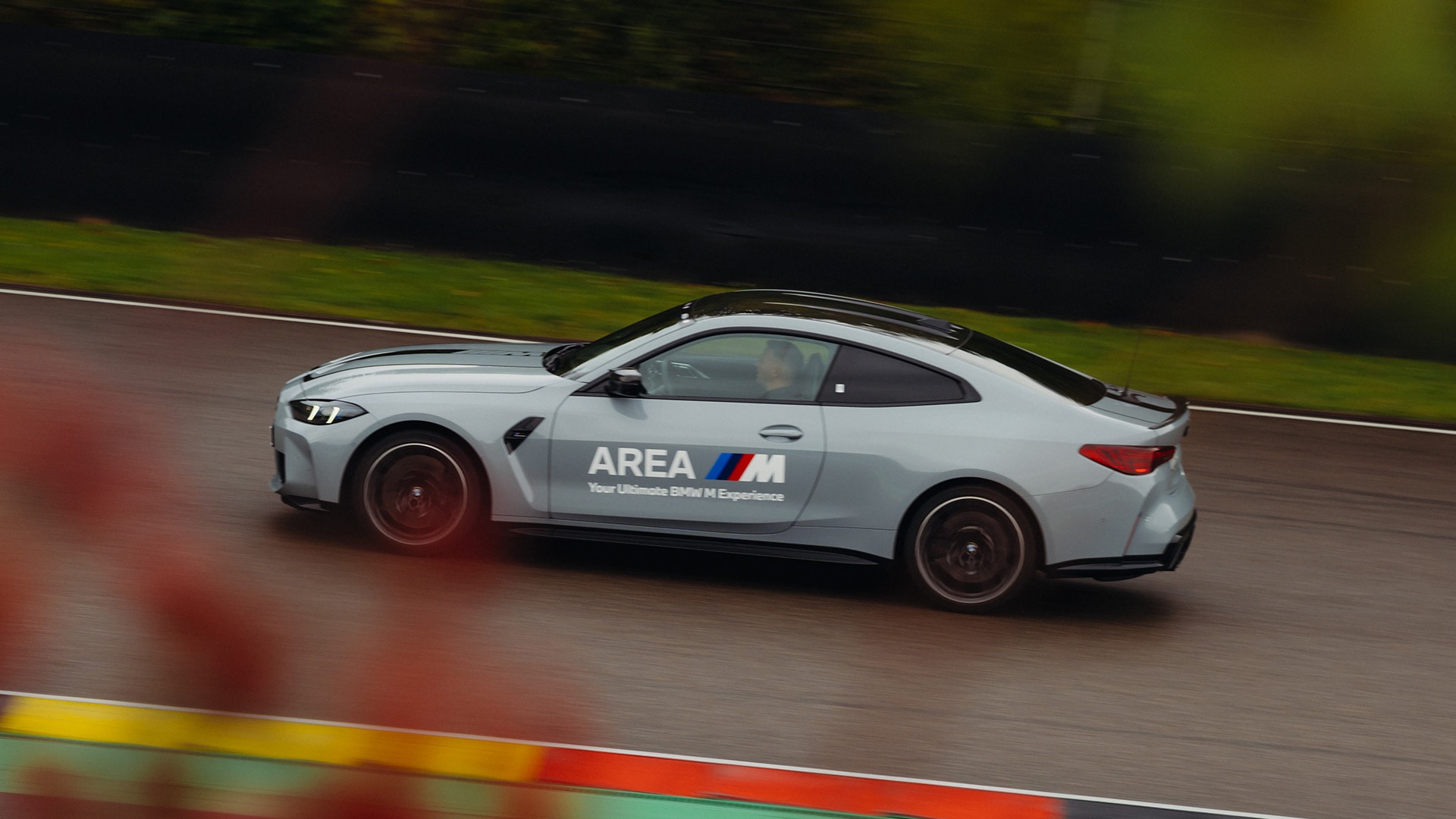 Ein silbener BMW M Sportwagen in Bewegung auf einer Rennstrecke. Das Fahrzeug trägt die Aufschrift 'AREA M' an der Seite mit dem Text 'Your Ultimate BMW M Experience'. Das Fahrzeug ist mit Bewegungsunschärfe vor einem dunkelgrünen Hintergrund aufgenommen, was auf hohe Geschwindigkeit auf der Asphaltbahn hindeutet.