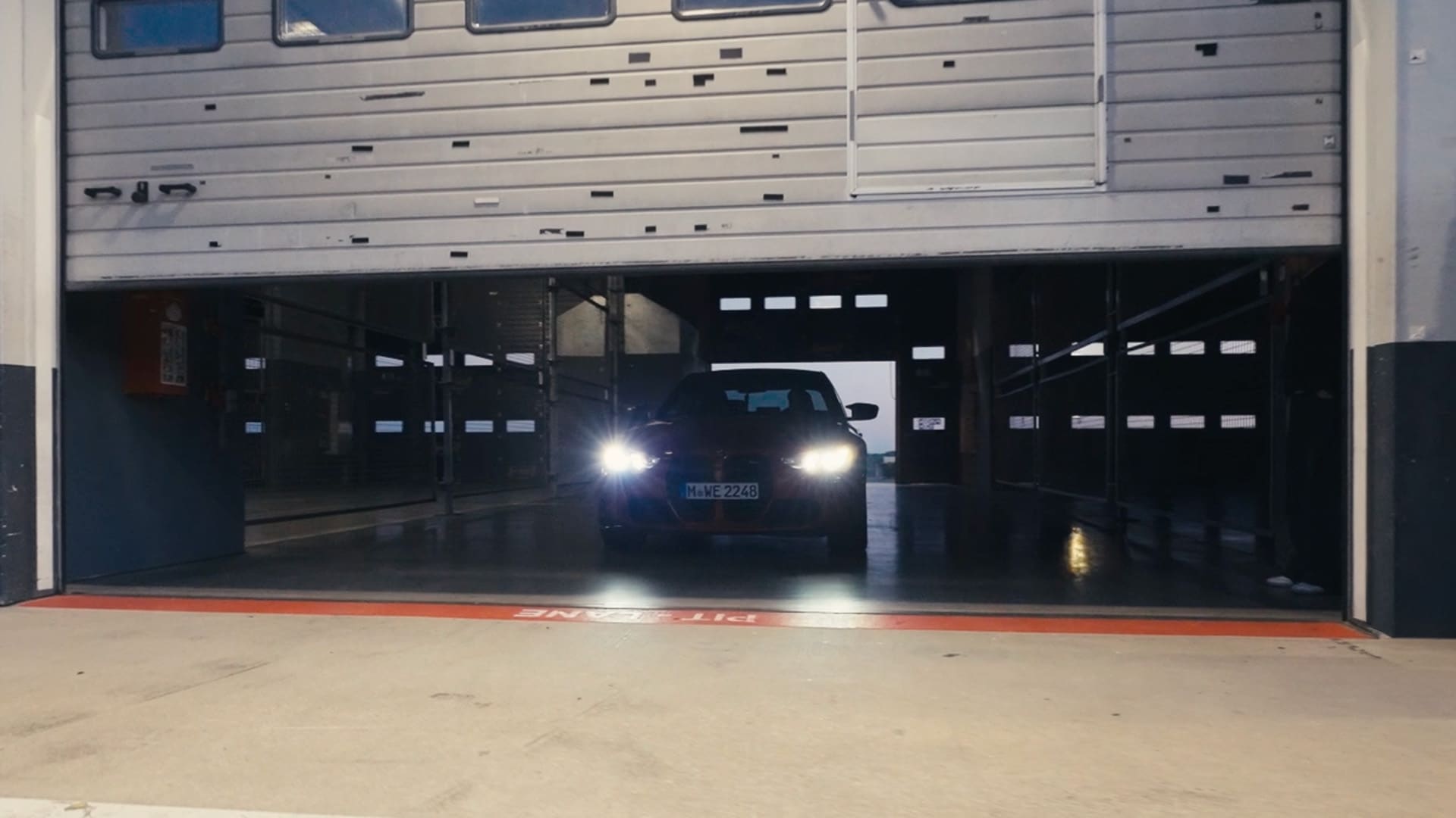 Ein dunkles Fahrzeug mit eingeschalteten Scheinwerfern verlässt eine Garage oder Werkstatt durch ein teilweise geöffnetes Rolltor. Das Kennzeichen des Fahrzeugs ist sichtbar, während es über eine rote Schwellenmarkierung am Eingang fährt.