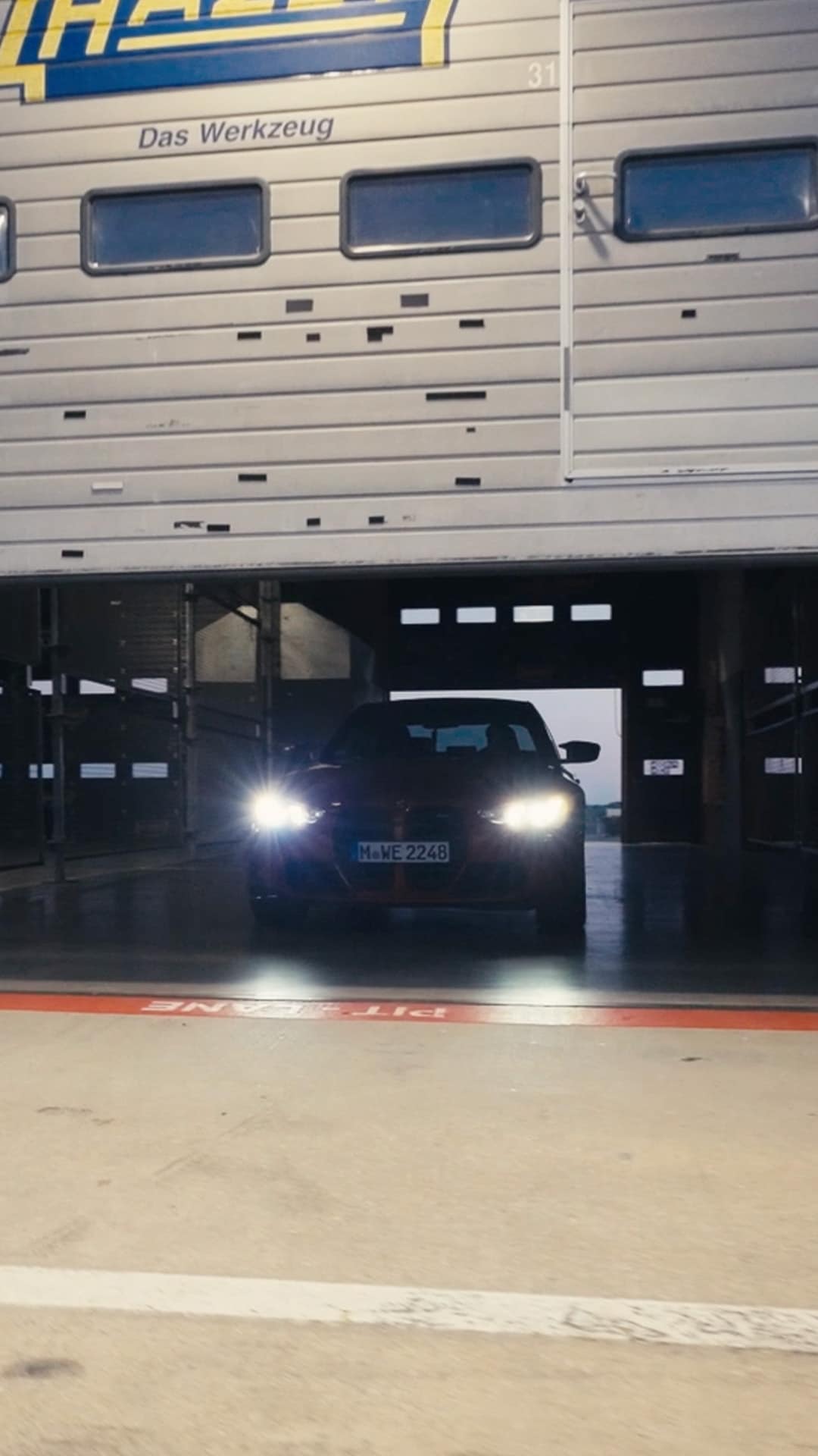 Ein dunkles Fahrzeug mit hellen Scheinwerfern fährt aus einer Garage oder Werkstatt heraus. Auf der Außenwand des Gebäudes steht 'Das Werkzeug' und es scheint Teil einer Anlage mit nummerierten Buchten zu sein.