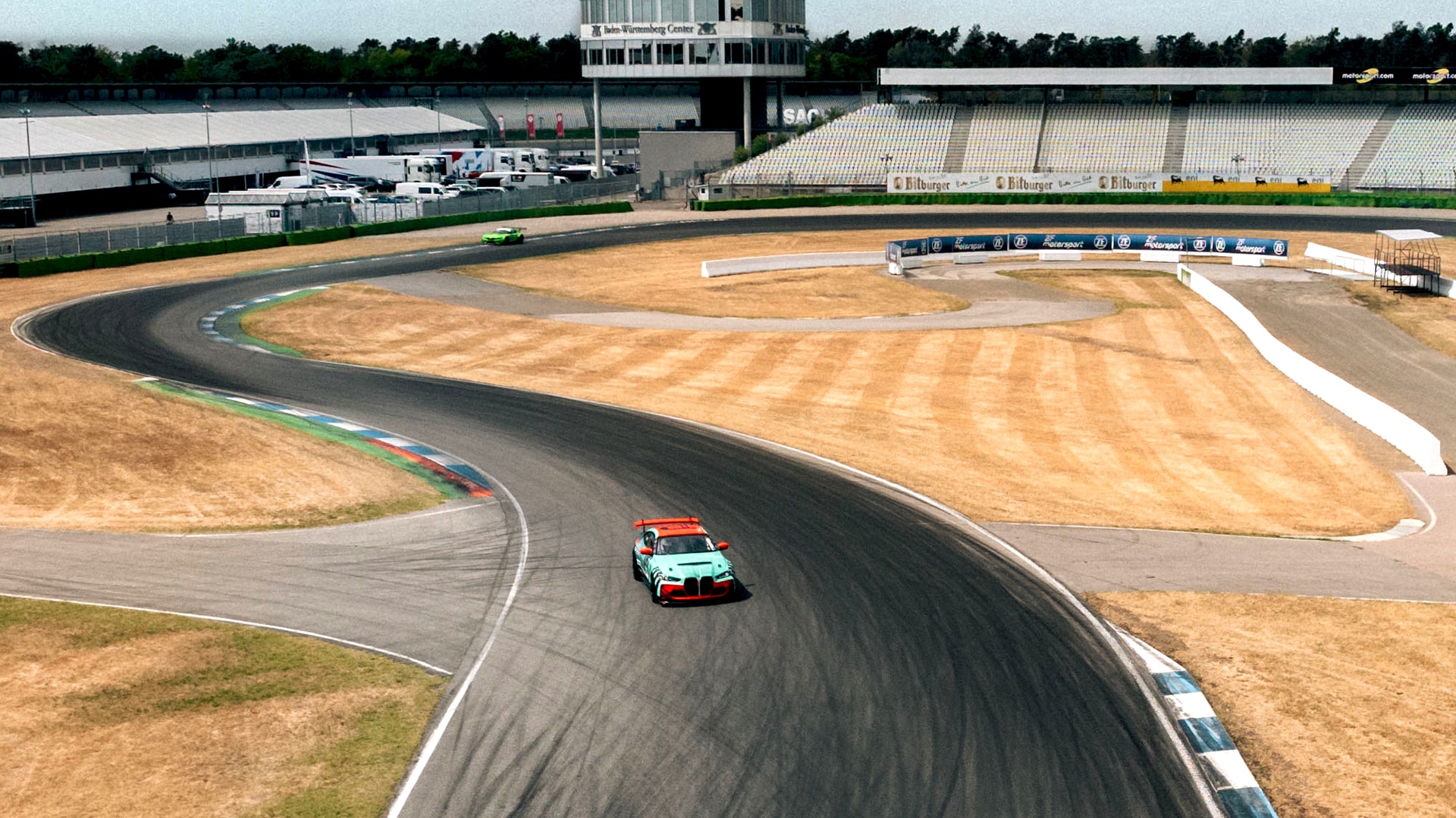 Eine Luftaufnahme einer Motorsport-Rennstrecke mit zwei Fahrzeugen auf der Strecke - einem rot-blauen Fahrzeug im Vordergrund und einem grünen Fahrzeug weiter hinten. Die Strecke weist mehrere Kurven mit Asphaltbelag auf, umgeben von sandigen Auslaufzonen. Im Hintergrund sind eine Zuschauertribüne und ein Kontrollturm zu sehen, sowie einige Servicefahrzeuge im Fahrerlager.