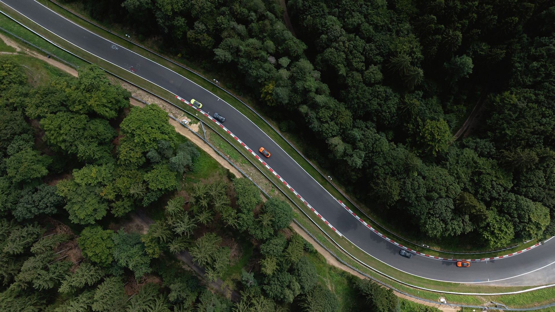 Luftaufnahme einer kurvenreichen Rennstrecke, die durch dichten Wald führt. Mehrere bunte Fahrzeuge sind zu sehen, wie sie den kurvigen Abschnitt der Strecke befahren, der mit rot-weißen Sicherheitsbarrieren an den Rändern versehen ist.