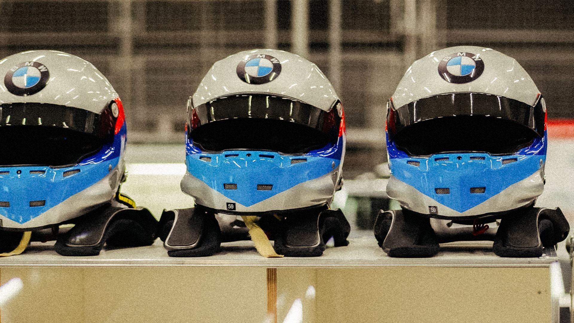 Drei Rennhelme mit BMW-Logos, die nebeneinander auf einem Regal ausgestellt sind. Jeder Helm hat ein charakteristisches blau-weiß-schwarzes Farbschema mit übereinstimmendem Design.