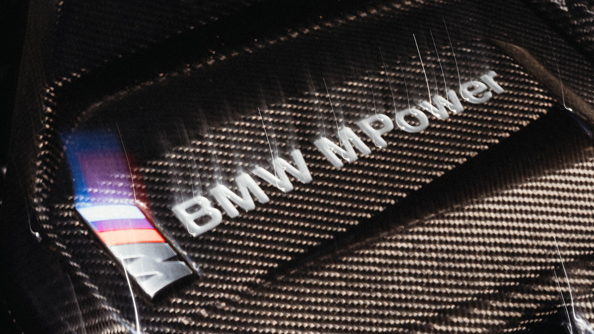 Nahaufnahme einer Kohlefaserkomponente mit dem BMW M Power Logo und dem charakteristischen BMW M Dreifarbemblem, eingebettet in die strukturierte Oberfläche.