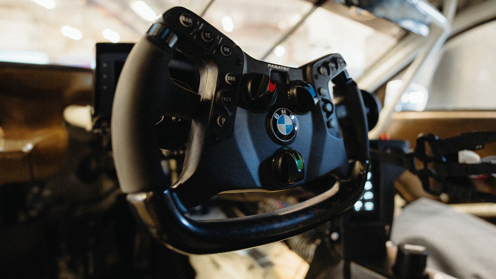Ein Fanatec-Rennsimulatoren-Lenkrad mit BMW-Branding. Das schwarze Lenkrad verfügt über mehrere Tasten und Bedienelemente auf der Vorderseite, einschließlich eines markanten BMW-Logos in der Mitte. Das Lenkrad ist an einem Rennsimulator-Gestell in einer Innenumgebung montiert.