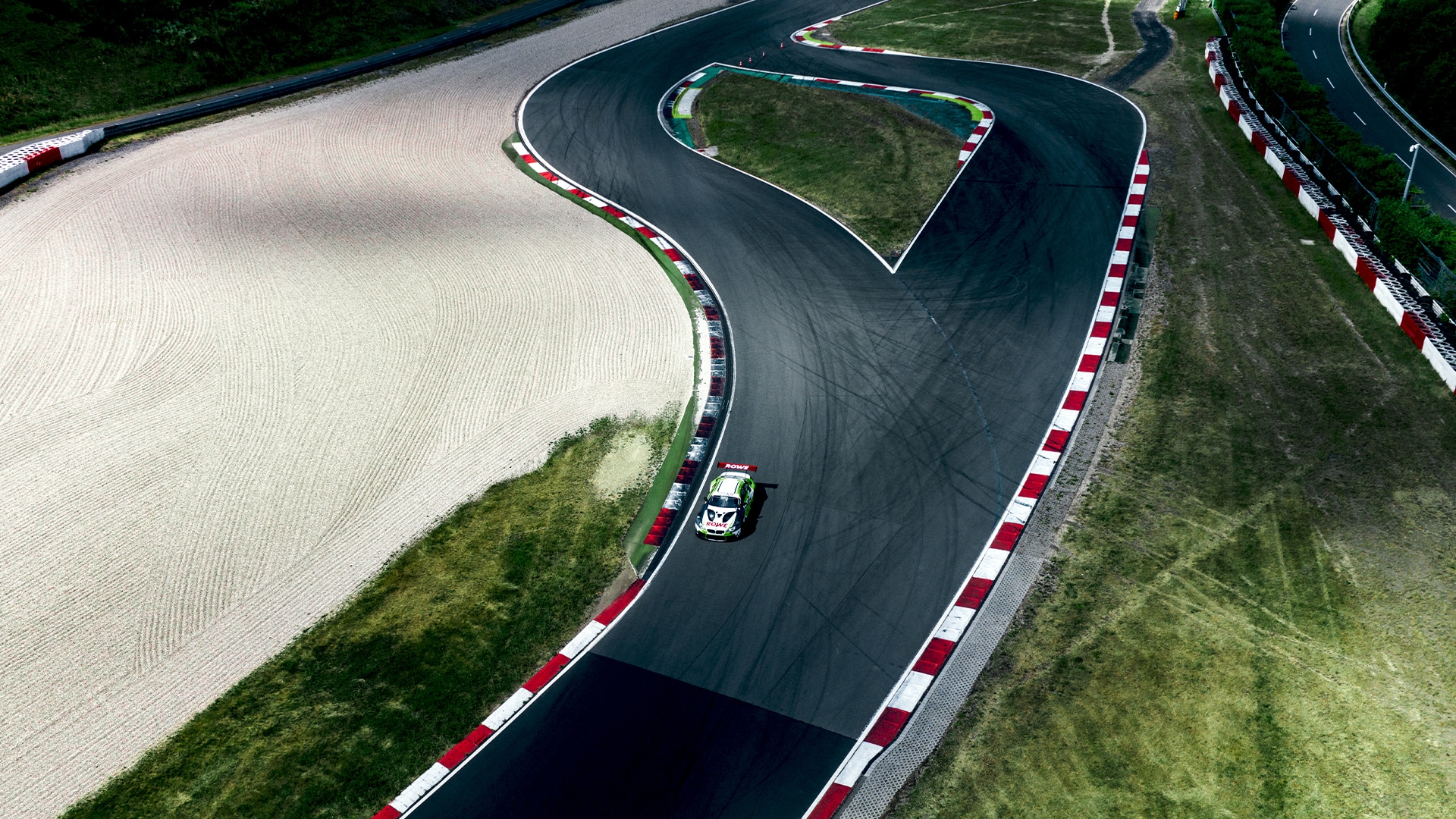 Luftaufnahme einer Rennstrecke mit einer scharfen S-Kurve, die von rot-weiß gestreiften Randsteinen begrenzt wird. Ein einzelnes Rennauto befährt die dunkle Asphaltpiste, die von Sandfallen und Grasflächen umgeben ist. Reifenspuren sind auf der Streckenoberfläche sichtbar.