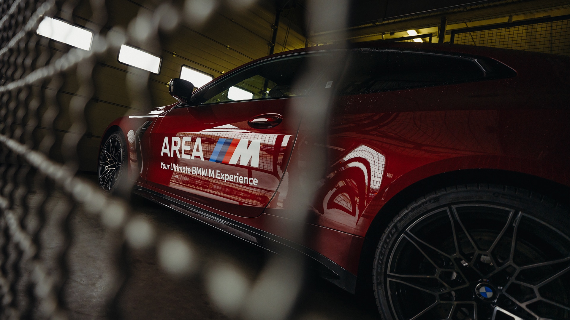 Ein glänzender roter BMW Sportwagen mit dem Text 'AREA M Your Ultimate BMW M Experience' auf der Seite, durch einen Metallzaun fotografiert. Das Fahrzeug steht in einer Innenanlage mit Deckenbeleuchtung.