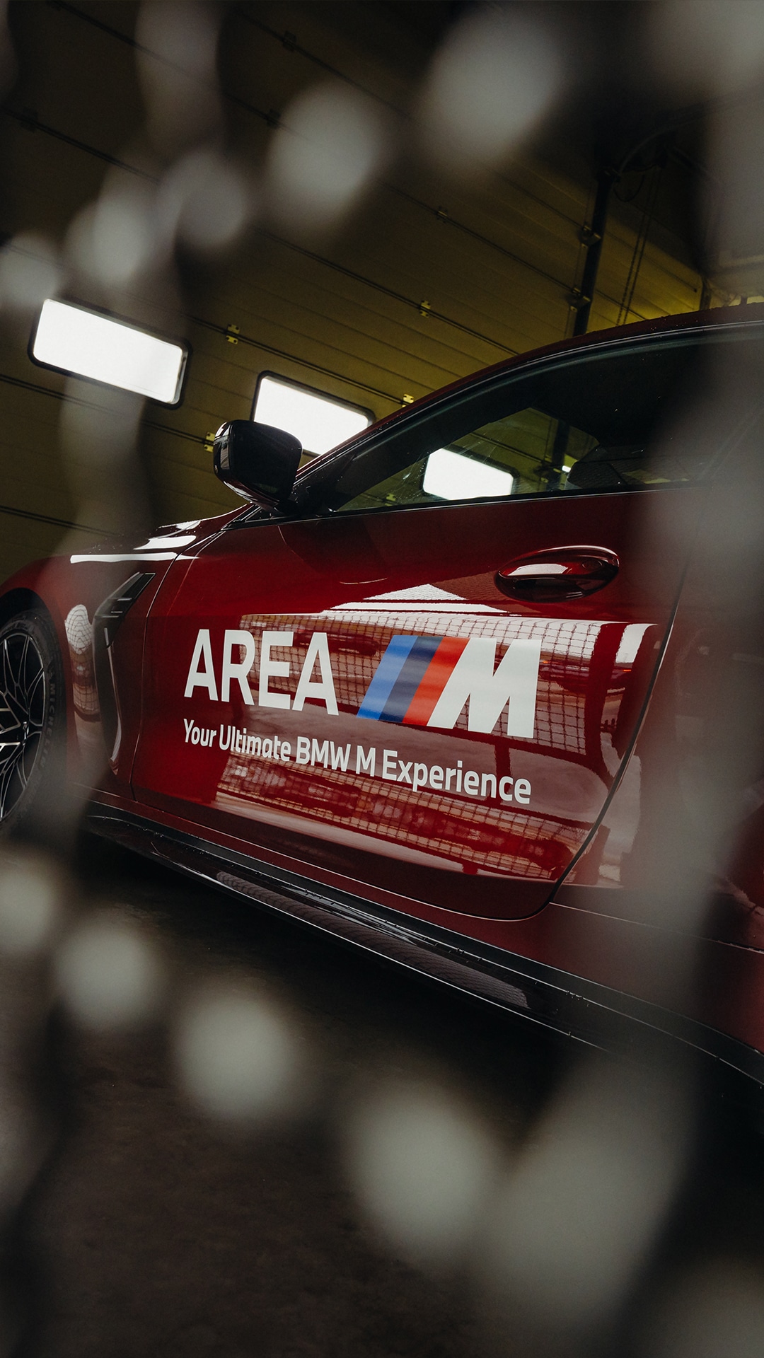 Ein roter BMW mit der Aufschrift 'AREA M' und 'Your Ultimate BMW M Experience' auf der Tür, fotografiert in einer Innenanlage mit sichtbaren Deckenleuchten. Das Bild wurde durch einen Zaun oder ein Gitter aufgenommen, was Bokeh-Effekte im Vordergrund erzeugt.