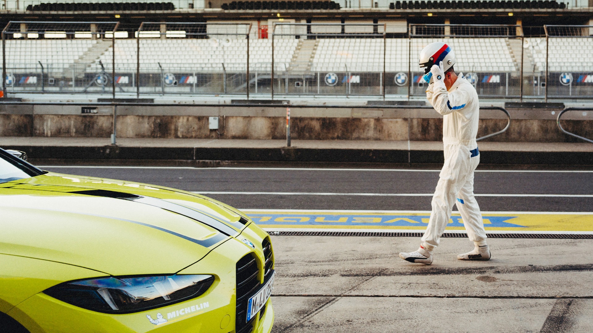 Eine Rennstrecke mit leeren Tribünen, die BMW M Branding zeigen. Im Vordergrund steht ein leuchtend gelber Sportwagen auf der Strecke, während eine Person in weißem Rennanzug und Helm entlang der Boxengasse geht. Die Szene zeigt die professionelle Motorsportumgebung mit deutlich sichtbaren Streckenmarkierungen.