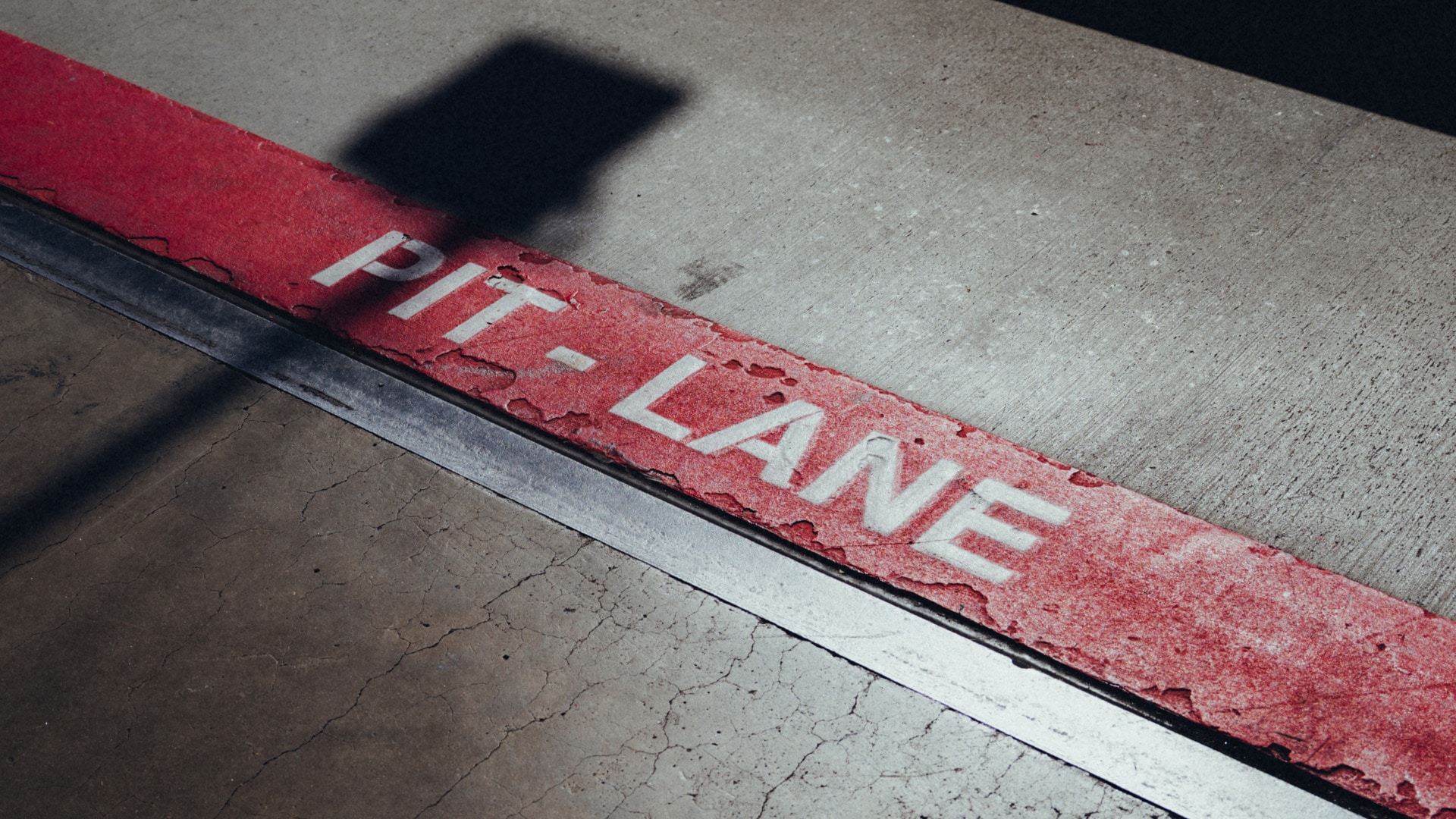 Eine rot gestrichene Linie auf einem Betonboden mit dem Text 'PIT LANE' in weißen Buchstaben. Ein Schatten fällt über einen Teil der Oberfläche.