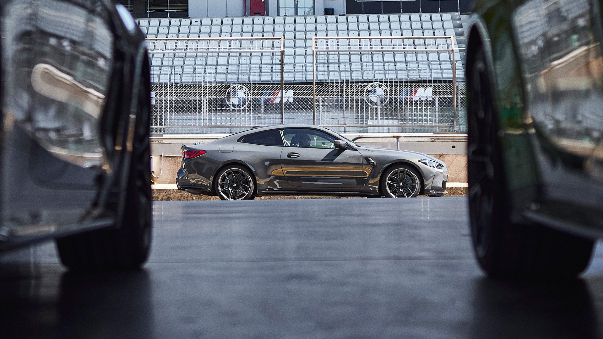 Ein elegantes graues BMW Sportcoupé, das auf einer Rennstrecke parkt, betrachtet durch eine Lücke zwischen zwei anderen Fahrzeugen. Im Hintergrund sind leere Tribünenplätze und Sicherheitsbarrieren zu sehen, mit BMW Logos auf den Streckenbarrieren.