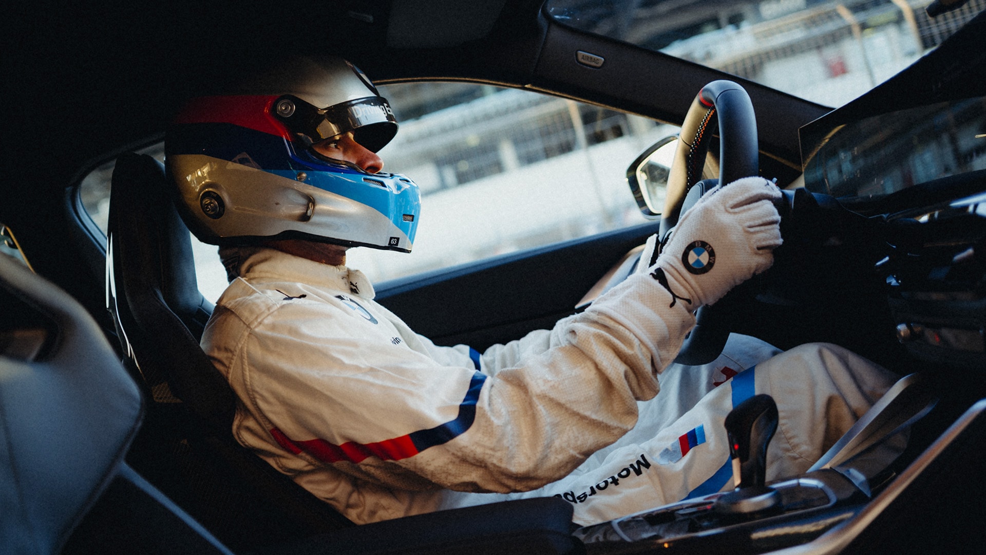 Ein Rennfahrer in einem weißen BMW Motorsport-Rennanzug und einem rot-blau-weißen Helm sitzt im Cockpit eines Fahrzeugs. Der Fahrer hält das Lenkrad mit weißen BMW-Rennhandschuhen fest. Das Interieur scheint zu einem Hochleistungsfahrzeug zu gehören, mit sichtbaren Rennsitzen.