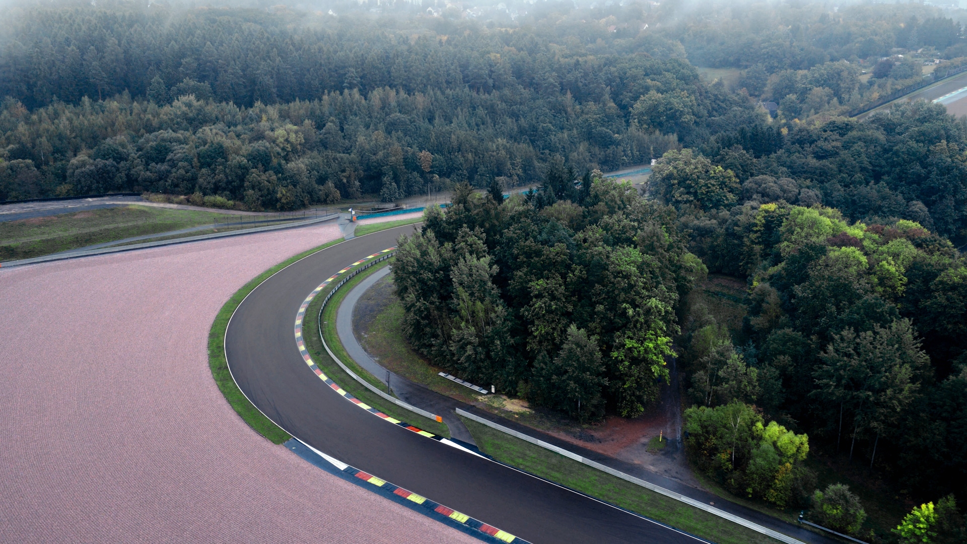 Luftaufnahme einer Rennstrecke mit einer gekrümmten Asphaltbahn mit farbigen Randsteinen, umgeben von dichten grünen Wäldern und Agrarflächen. Ein Fluss schlängelt sich in der Landschaft im Hintergrund.