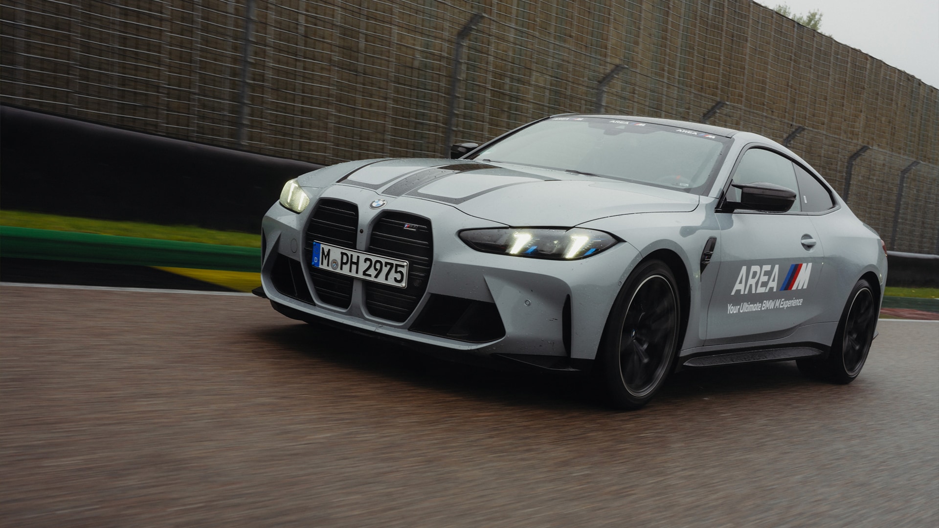 Ein silberner BMW M Sportwagen fährt auf einer Rennstrecke. Das Fahrzeug zeigt den markanten großen BMW-Kühlergrill und trägt die Aufschrift 'AREA M' an der Seite. Es wurde in Bewegung fotografiert, mit einer Schallschutzmauer im Hintergrund.