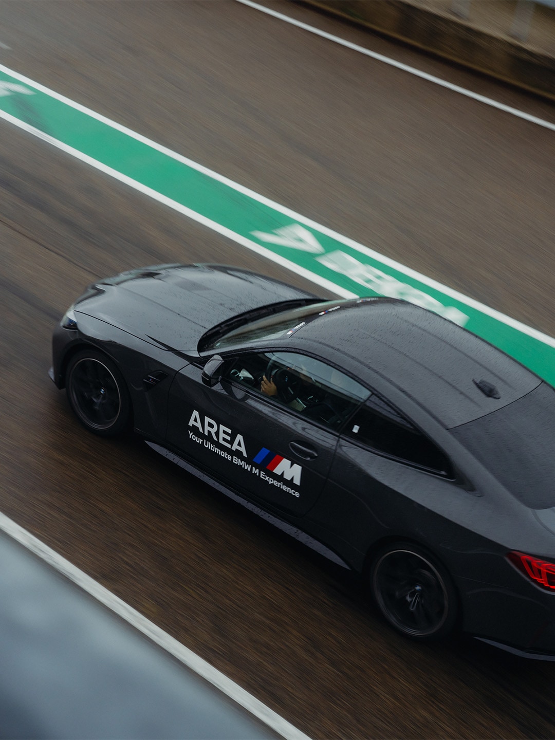 A black BMW M performance car driving on a race track with green and white lane markings. The vehicle displays 'AREA' and 'Your Ultimate BMW M Experience' text on its side, with the BMW M logo and tricolor stripe visible.