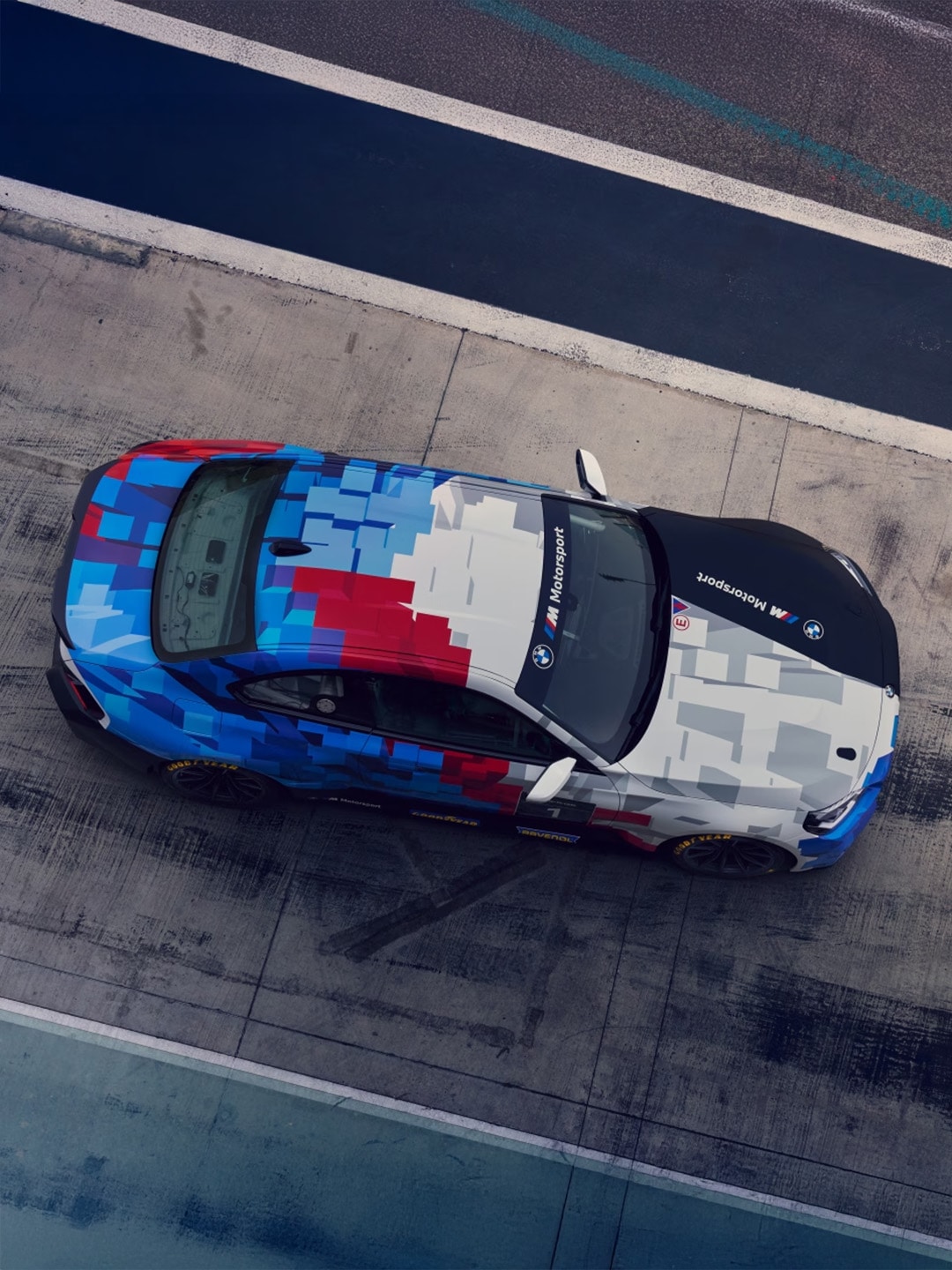 Overhead view of a BMW M sport car with a pixelated livery in blue, red, and white colors parked on a concrete surface next to a dark blue water feature or pool.