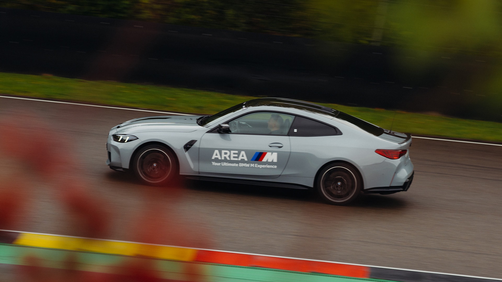 A grey BMW M performance car with 'AREA M' branding on its side drives on a race track. The vehicle is captured in motion with motion blur in the background, showing green grass and trees. Colorful track markings in red, yellow, and green are visible at the bottom of the image.