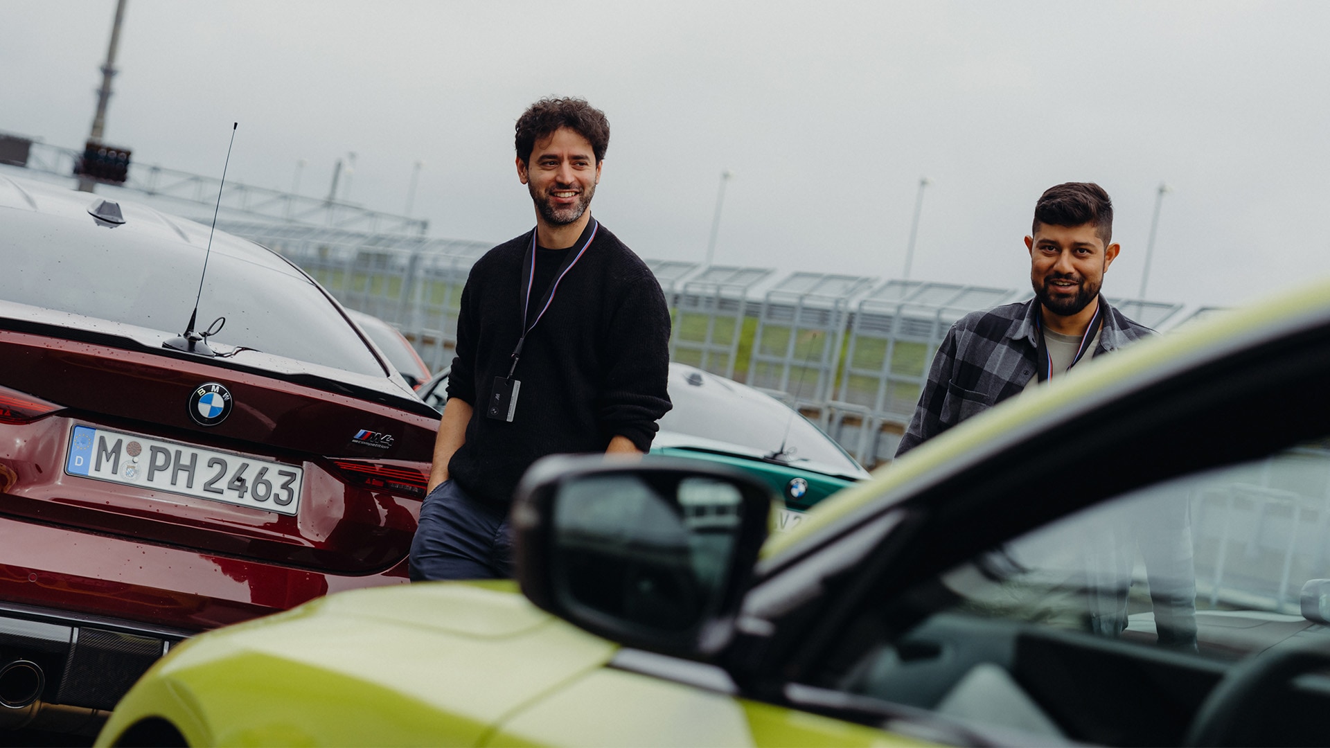 Two men stand between parked cars. The man on the left wears a black jacket and lanyard, smiling at the camera. The man on the right wears a checkered shirt and also smiles. A red BMW with license plate M-PH2463 is visible on the left, with a yellow car in the foreground. Multiple vehicles and greenhouses are blurred in the background under an overcast sky.