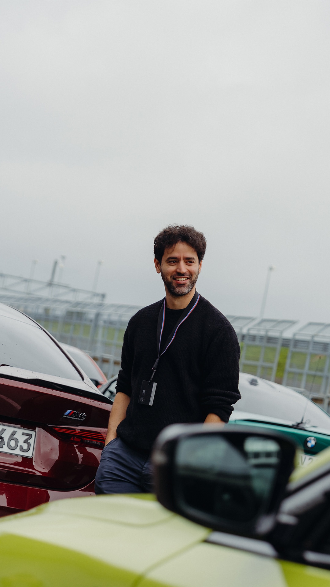 A smiling man with dark curly hair and a beard wearing a black sweater and ID badge stands next to a red car in a parking lot. Multiple vehicles are visible in the background with an overcast sky and industrial buildings.