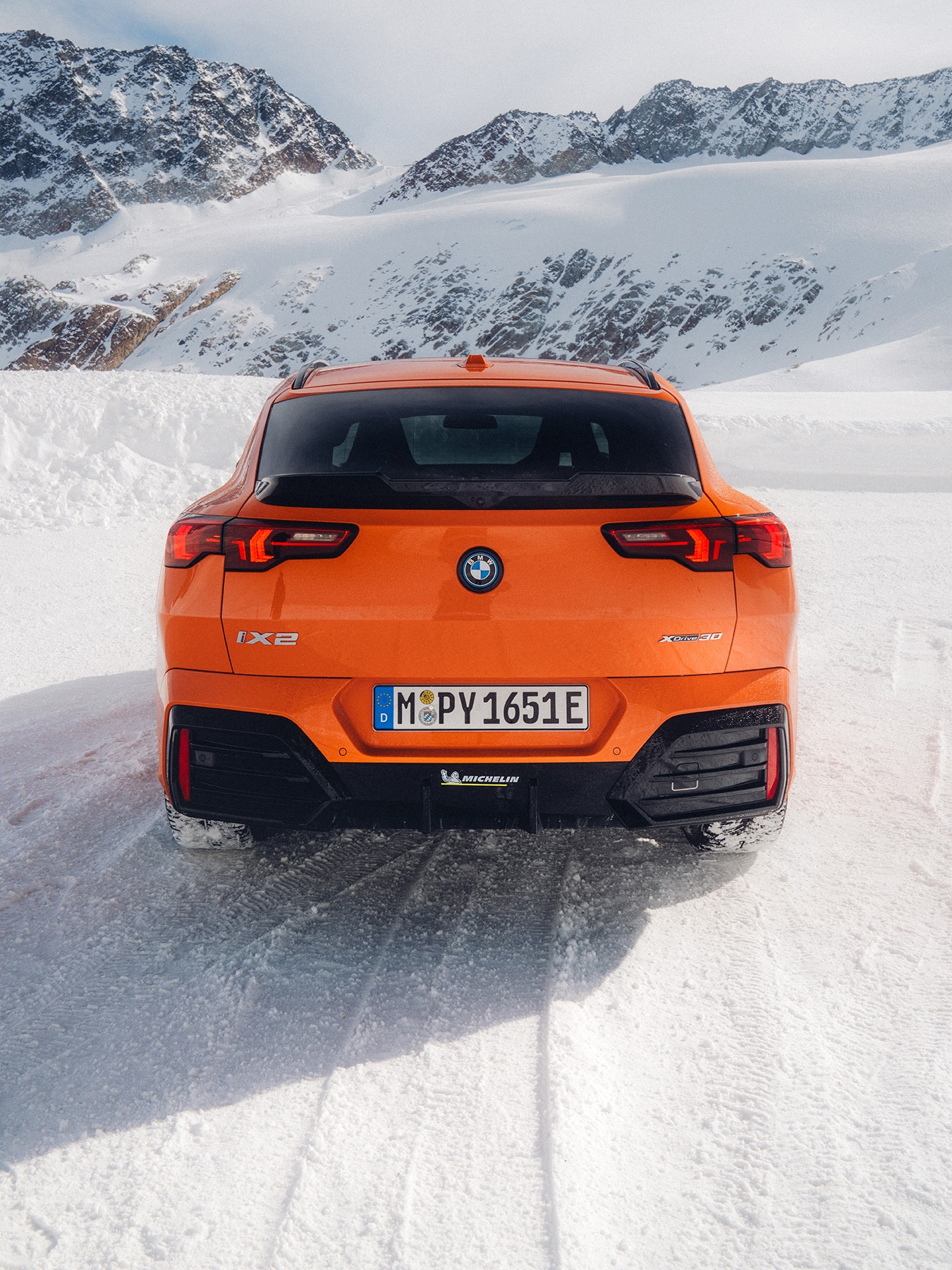 Rückansicht eines orangefarbenen BMW iX2 xDrive30 Elektrofahrzeugs, das in einer schneebedeckten Landschaft mit schneebedeckten Bergen im Hintergrund parkt. Das Auto zeigt ein deutsches Kennzeichen und Michelin-Branding.