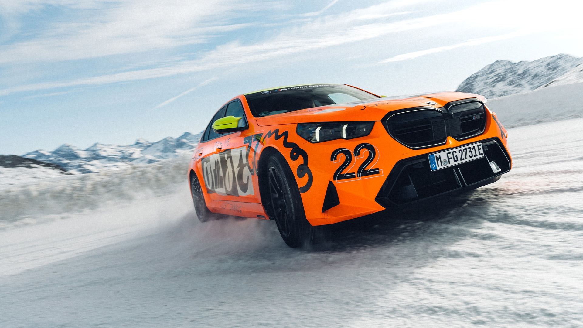Ein leuchtend orangefarbener Sportwagen mit der deutlich sichtbaren Nummer 22 an der Seite driftet über eine verschneite Landschaft. Das Fahrzeug, anscheinend ein BMW mit deutschem Kennzeichen, wirbelt Schnee auf, während es durch eine winterliche Berglandschaft fährt. Im Hintergrund sind schneebedeckte Gipfel unter einem blauen Himmel mit dünnen Wolken zu sehen.