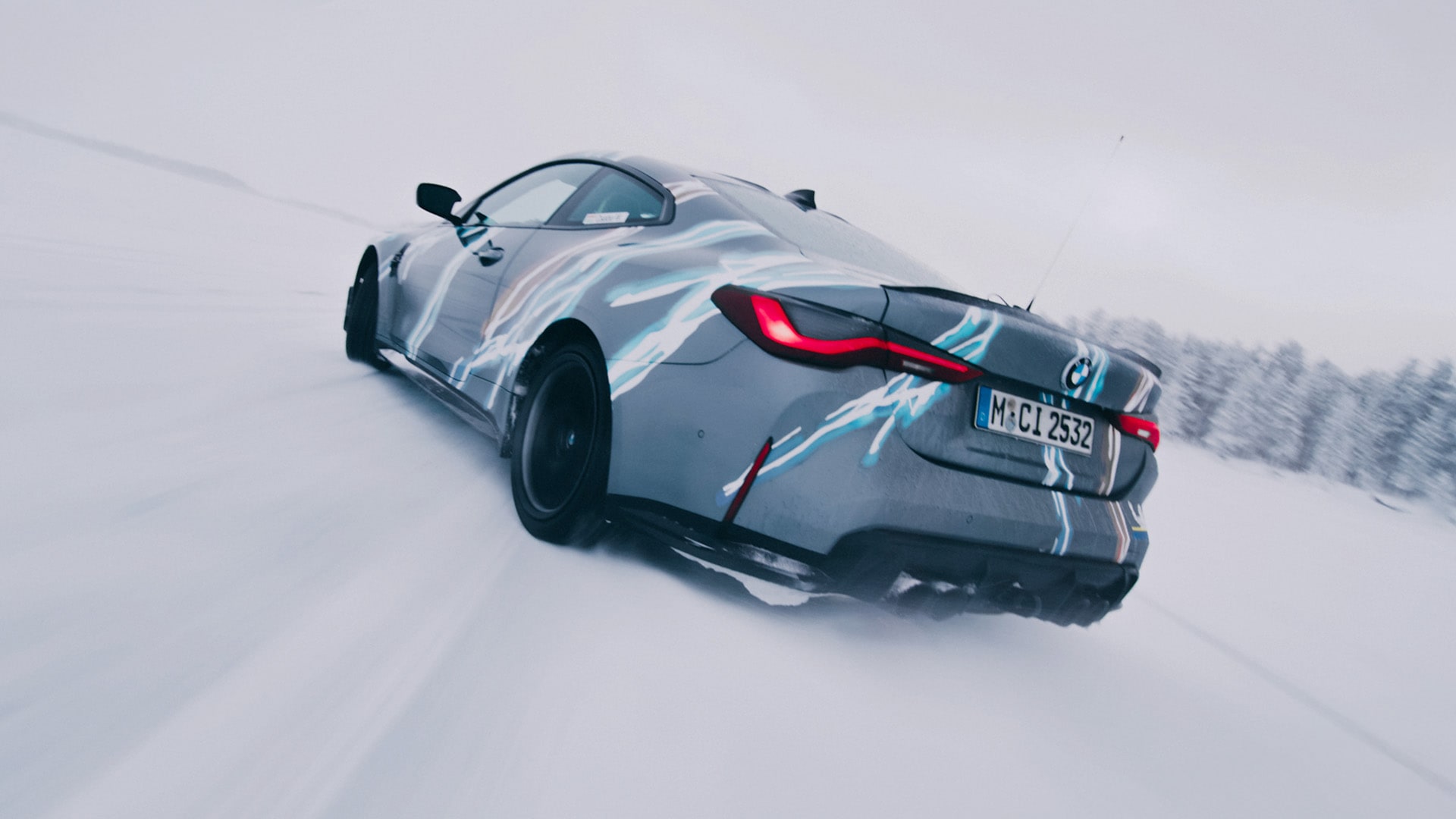 Ein BMW Sportwagen mit Tarnfolie fährt durch Schnee. Das Fahrzeug, erkennbar an seinen markanten roten Rücklichtern und Münchner Kennzeichen, hinterlässt Spuren im weißen, schneebedeckten Gelände mit verschwommenen Bäumen im Hintergrund.