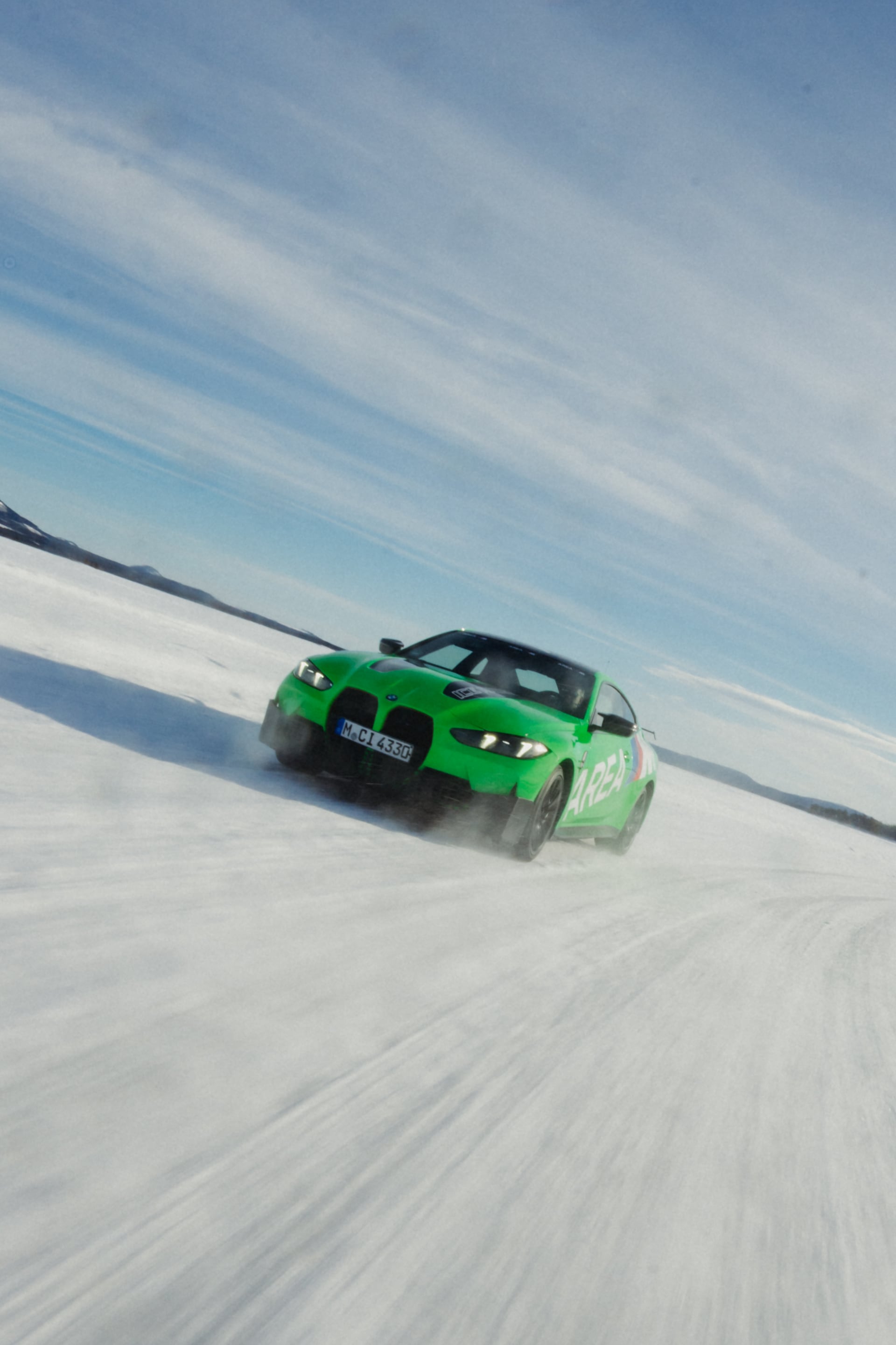 A bright green sports car driving across a snowy frozen landscape under a clear blue sky with wispy clouds. Snow sprays from the vehicle's tires as it moves across the white terrain.