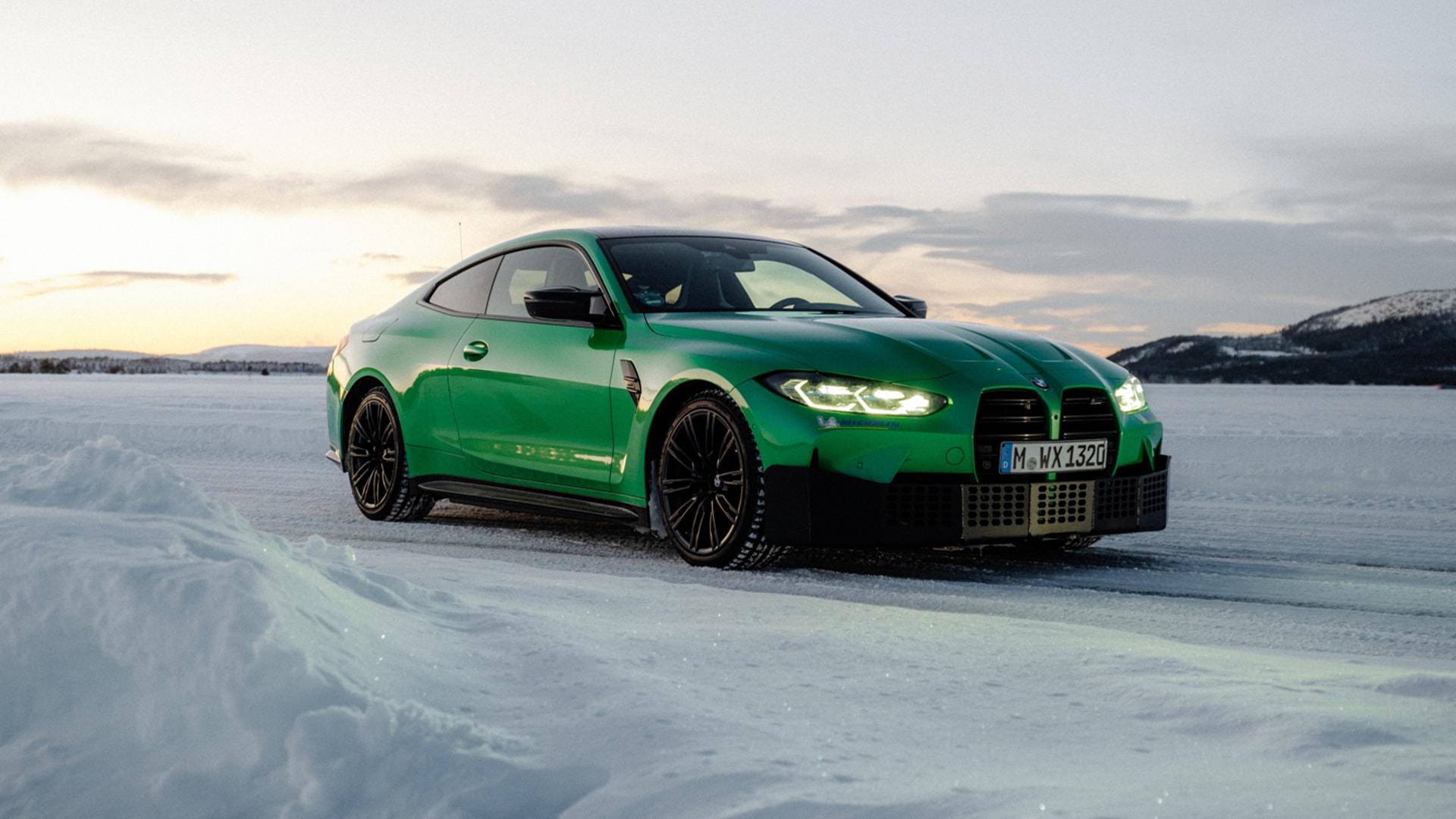 Ein leuchtend grüner BMW M4 Sportwagen, der auf einer verschneiten Fläche parkt, mit eingeschalteten Scheinwerfern. Die Szene zeigt eine Winterlandschaft mit schneebedecktem Boden, Bergen im Hintergrund und einem bewölkten Himmel in der Dämmerung.