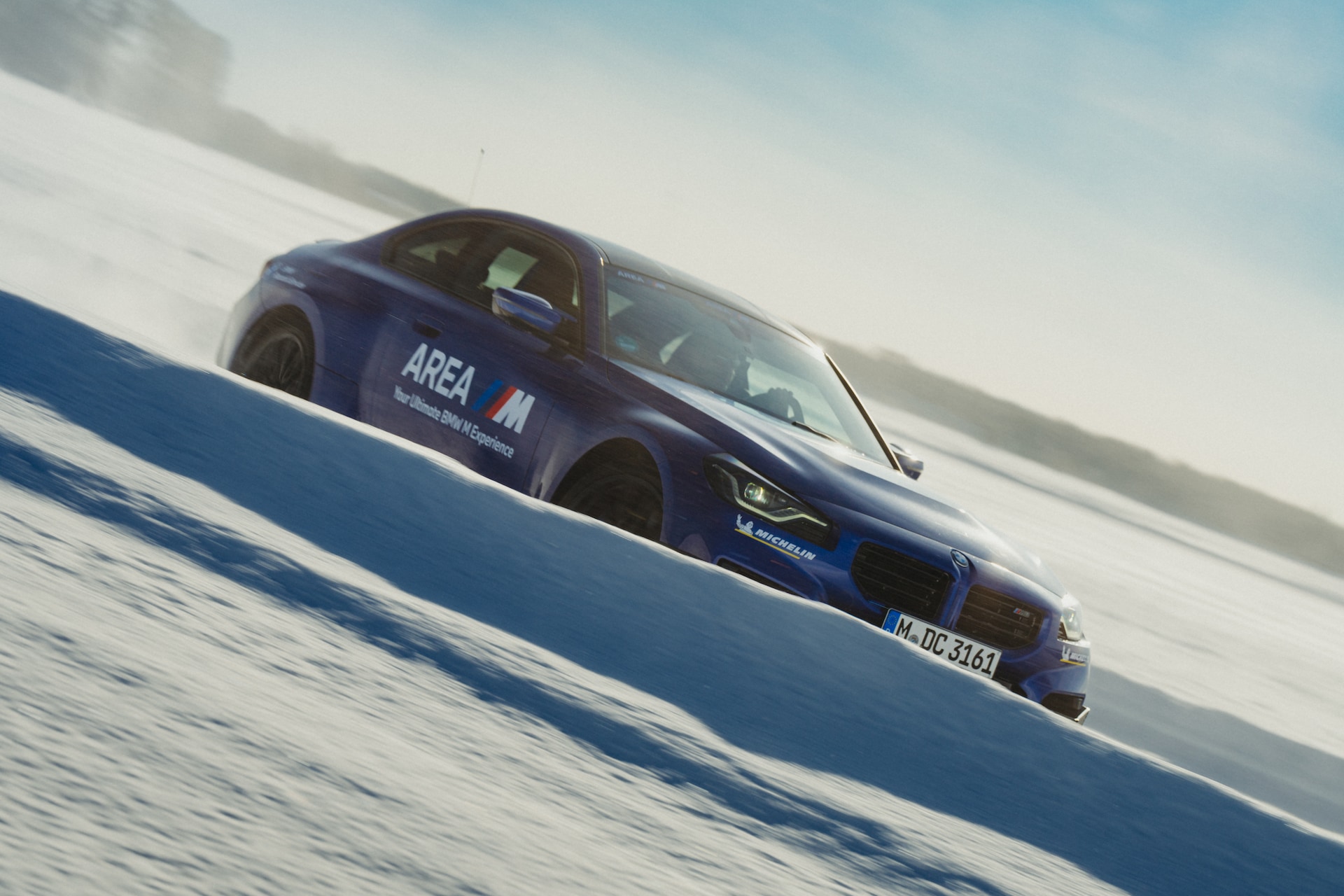 Ein dunkelblaues BMW Fahrzeug mit 'AREA'-Branding und Michelin-Reifen fährt diagonal über einen verschneiten Berghang. Das Fahrzeug wird in Bewegung vor einem hellen, bewölkten Himmel mit schneebedecktem Gelände im Hintergrund erfasst.