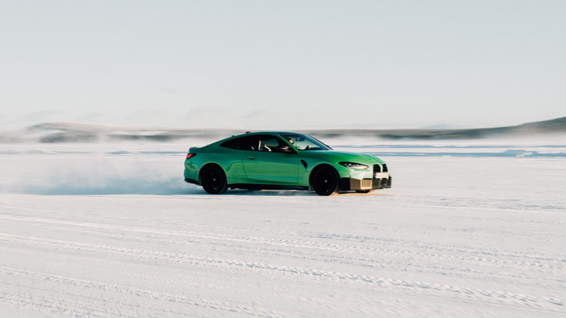 Ein leuchtend grüner BMW Sportwagen, der über eine verschneite Landschaft driftet und dabei eine Schneewolke um sich herum erzeugt. Das Fahrzeug hat schwarze Räder und ist vor einem blassen Himmel auf einer schneebedeckten Oberfläche mit sichtbaren Reifenspuren fotografiert.