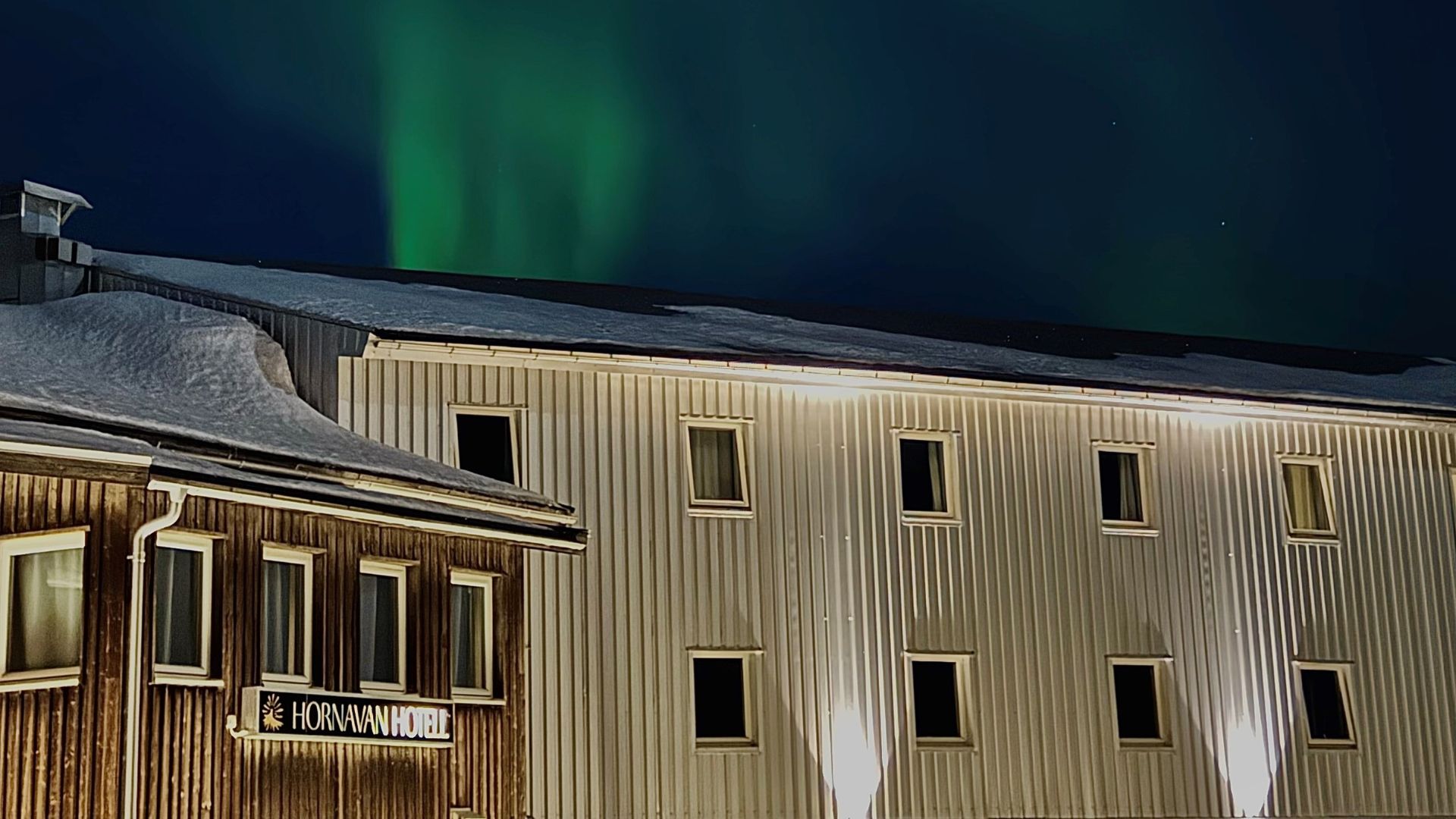 Eine Nachtansicht des Hornavan Hotels mit schneebedeckten Dächern. Der dunkle Himmel darüber zeigt lebhafte grüne Nordlichter (Aurora Borealis). Das Gebäude hat einen hölzernen braunen Abschnitt auf der linken Seite mit einem Hotelschild und einen größeren, helleren Metallabschnitt auf der rechten Seite mit mehreren Fenstern.