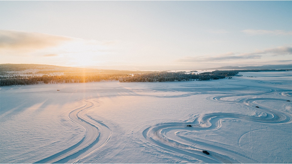 Luftaufnahme einer schneebedeckten Landschaft mit gewundenen Spuren oder Pfaden im Schnee. Die Sonne geht am Horizont unter und wirft einen goldenen Schimmer über die Szene. In der Ferne sind bewaldete Hügel und Berge unter einem klaren blauen Himmel mit vereinzelten Wolken zu sehen.