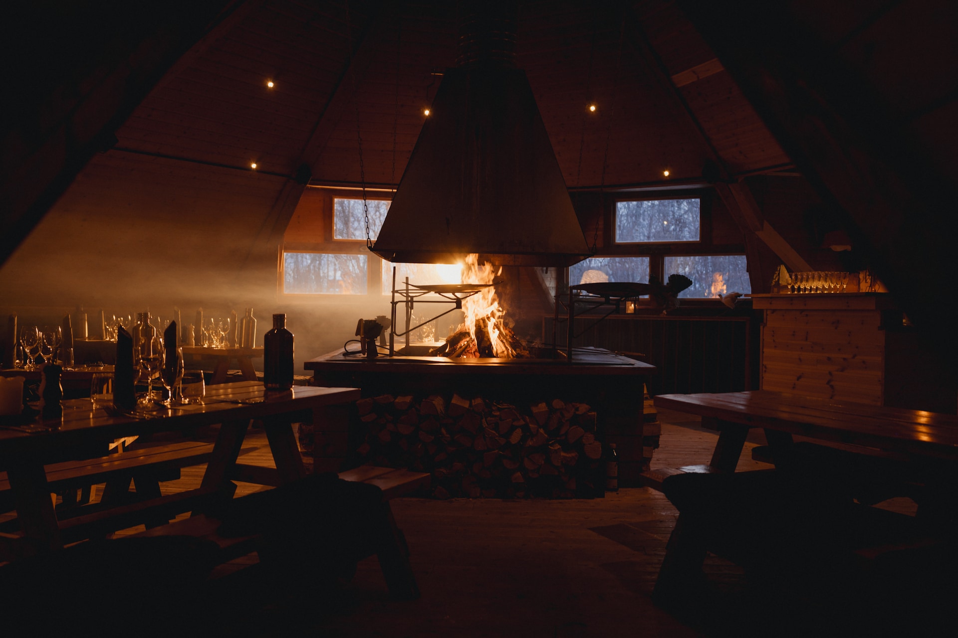Innenraum einer rustikalen Holzhütte bei Nacht mit einem großen zentralen Kamin, der hell brennt. Der Raum verfügt über Holzbänke, warme Umgebungsbeleuchtung von Deckenleuchten, große Fenster mit verschneiter Landschaft außen und verschiedene Flaschen sowie Gegenstände auf Tischen. Rauch steigt vom Feuer auf und beleuchtet die hölzerne Kassettendecke.