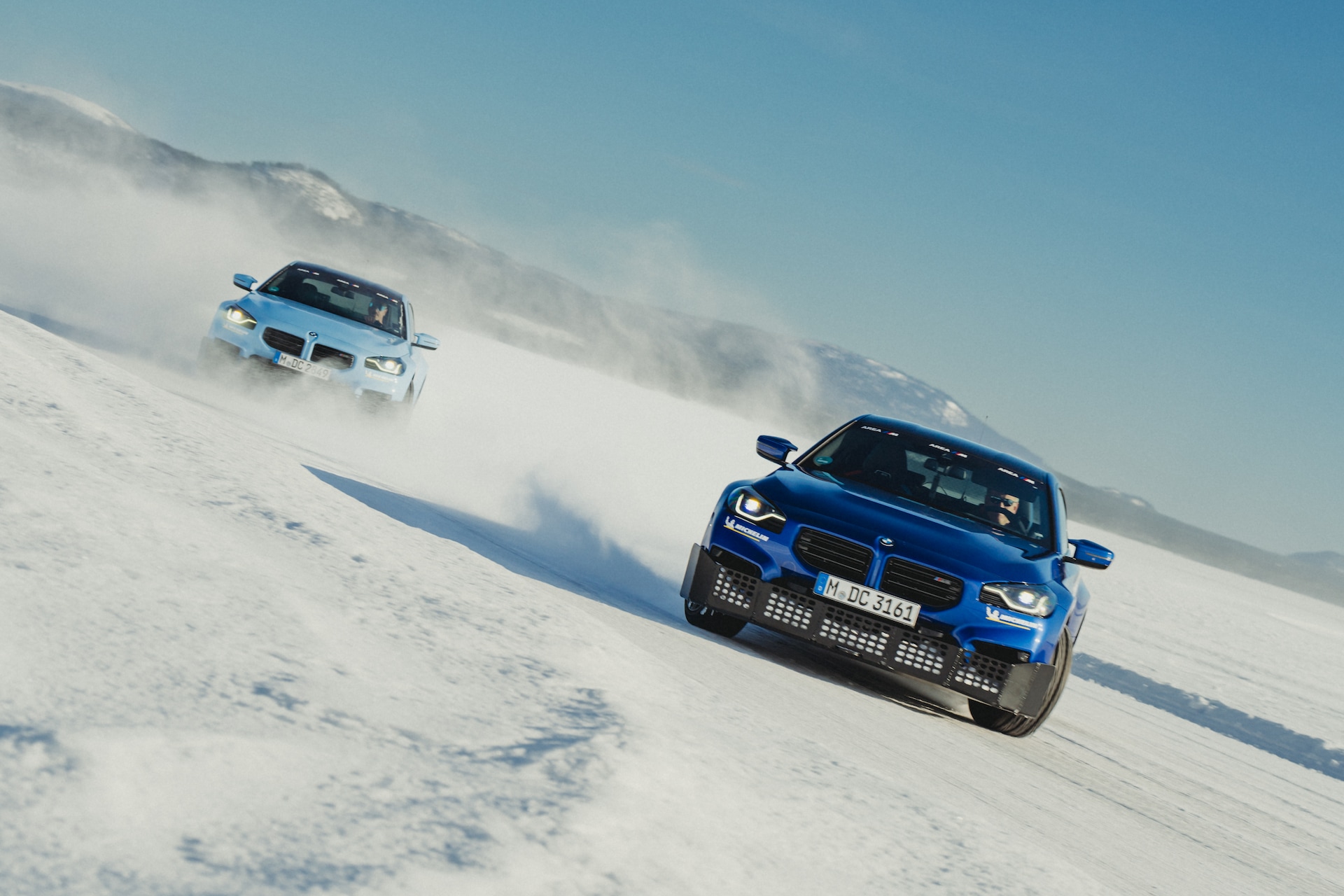Zwei blaue BMW Fahrzeuge fahren einen verschneiten Berghang hinunter und wirbeln Schneewolken auf. Das vordere Auto ist näher an der Kamera, während ein zweites Auto dahinter folgt. Schneebedeckte Berge sind im Hintergrund unter einem klaren blauen Himmel sichtbar.