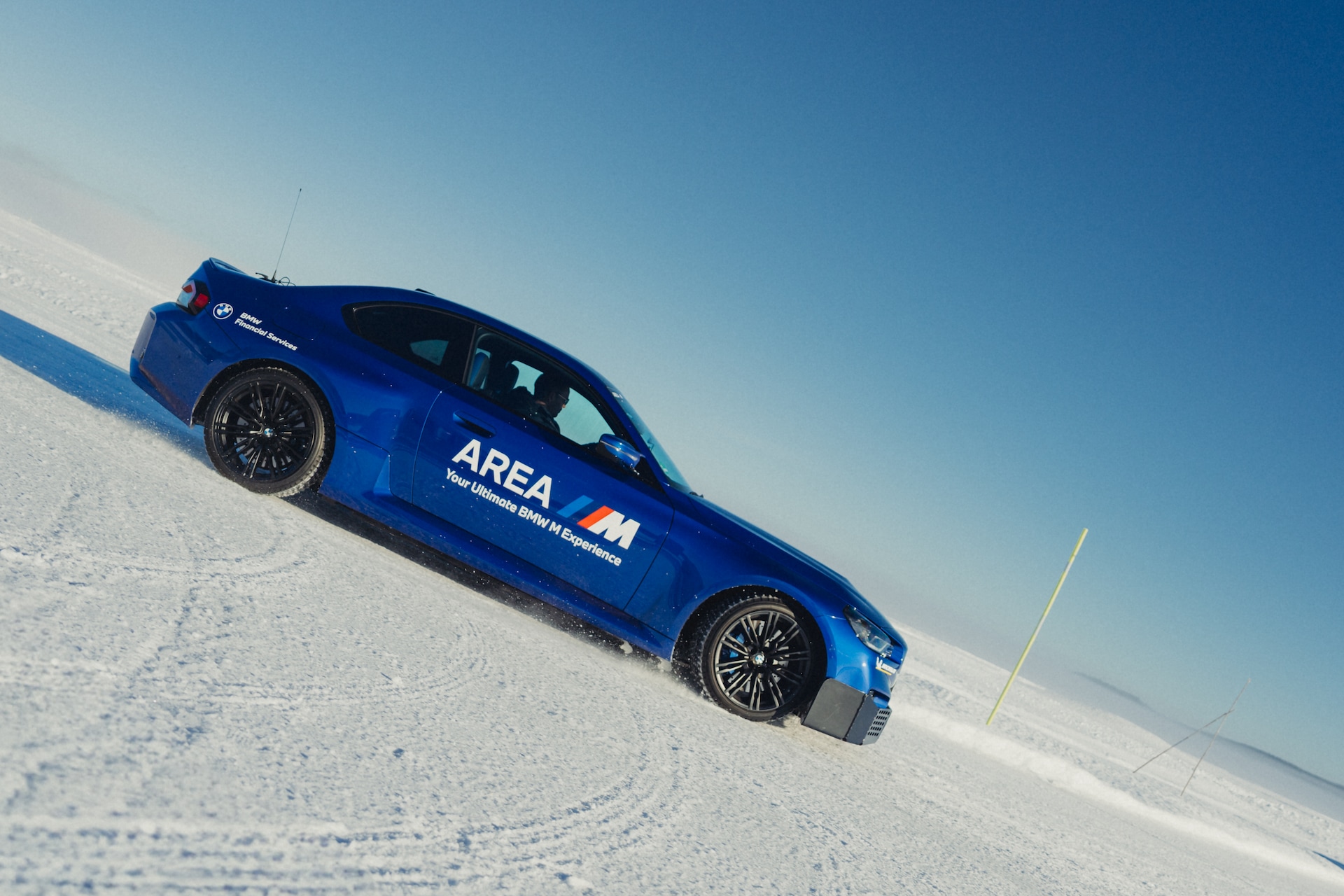 Ein leuchtend blaues BMW M Leistungsfahrzeug mit 'AREA M' Beschriftung fährt über eine verschneite Landschaft unter klarem blauen Himmel. Das Fahrzeug befindet sich auf leicht geneigtem Gelände mit schneebedecktem Terrain und einer gelben Markierungsstange im Hintergrund.