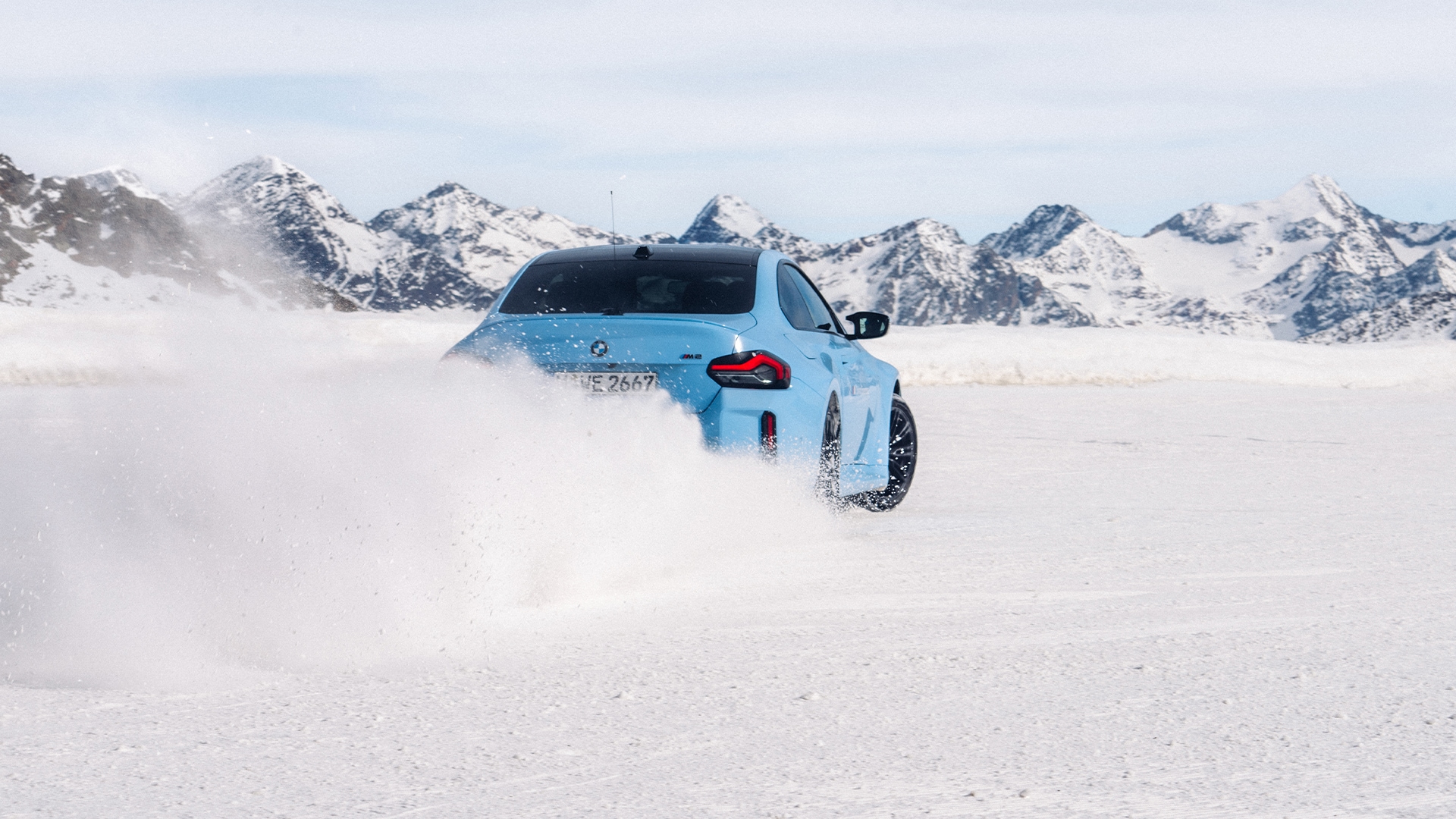 Ein hellblauer BMW fährt durch Schnee und wirbelt Pulverschnee auf, während es sich über eine weiße Schneelandschaft bewegt. Im Hintergrund sind schneebedeckte Berge unter einem bewölkten Himmel zu sehen.