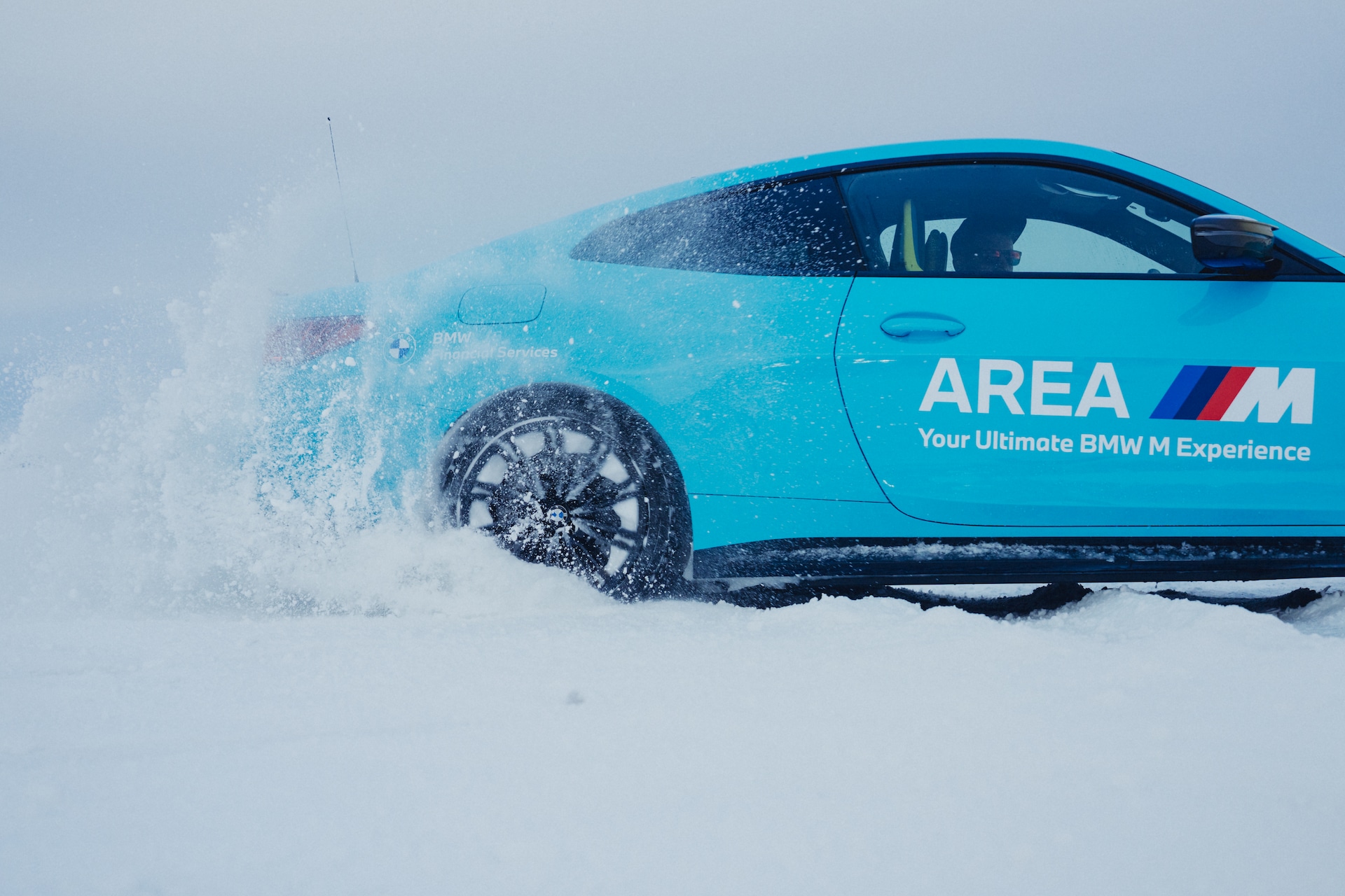 Ein leuchtend türkisfarbenes BMW Fahrzeug mit 'AREA M' Branding und dem Text 'Your Ultimate BMW M Experience' auf der Seite, das durch tiefem Schnee fährt. Schnee spritzt dramatisch von den Rädern und bedeckt die Windschutzscheibe, während das Fahrzeug durch eine verschneite Landschaft fährt.