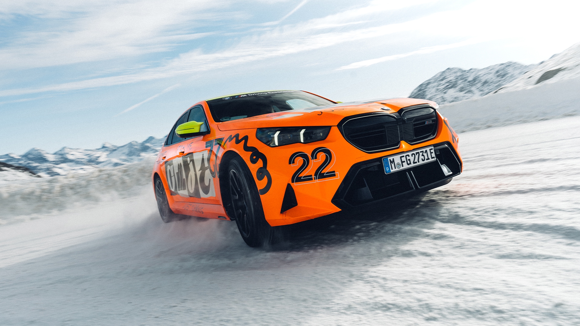 Ein leuchtend oranger M5 mit der Nummer 22 und dekorativen Grafiken fährt durch eine verschneite Landschaft mit Bergen im Hintergrund unter einem hellblauen Himmel mit dünnen Wolken.