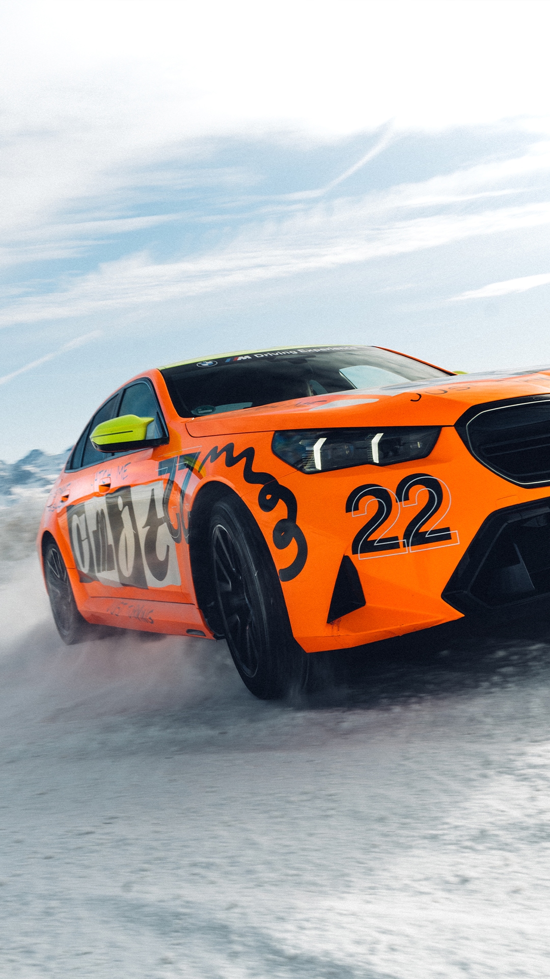 Ein leuchtend oranger M5 mit der Nummer 22 und dekorativen Grafiken, der über eine verschneite Oberfläche fährt. Das Auto hat neongrüne Seitenspiegel und wirbelt Schnee auf, während es sich unter einem hellblauen Himmel mit dünnen Wolken bewegt.