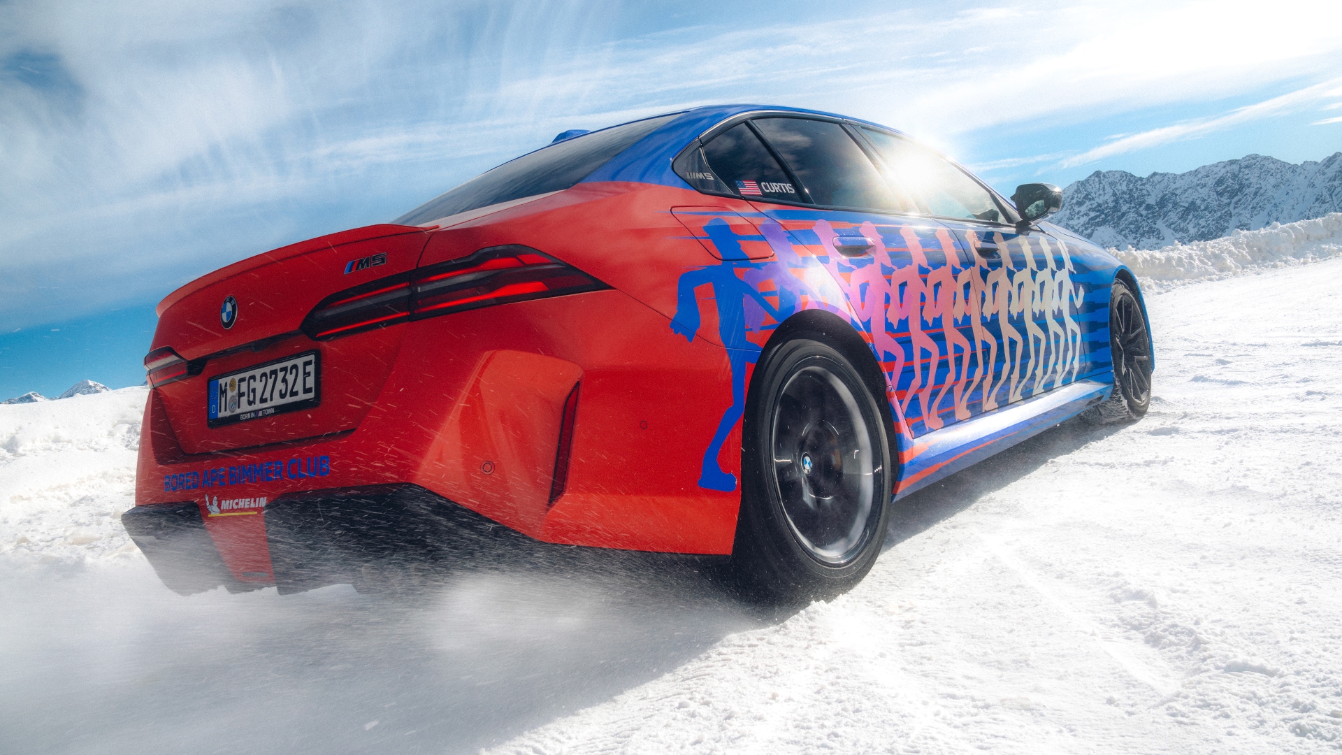 Ein roter BMW Sportwagen mit blauen grafischen Mustern an der Seite, der in einer verschneiten Landschaft parkt. Das Fahrzeug hat ein deutsches Kennzeichen und steht vor einem Hintergrund aus schneebedeckten Bergen unter blauem Himmel.