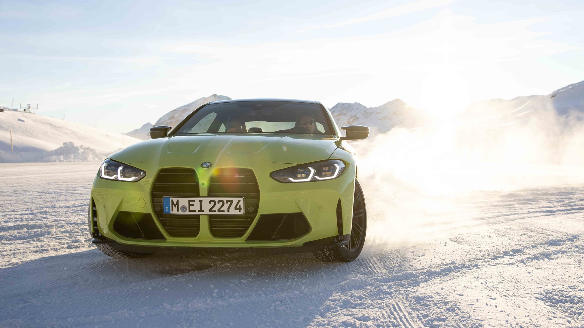 Ein leuchtend grüner BMW Sportwagen mit dem Kennzeichen M-EI 2274 fährt auf einer verschneiten Oberfläche. Das Fahrzeug wirbelt Schnee auf während es sich bewegt, mit schneebedeckten Bergen im Hintergrund unter einem hellblauen Himmel.