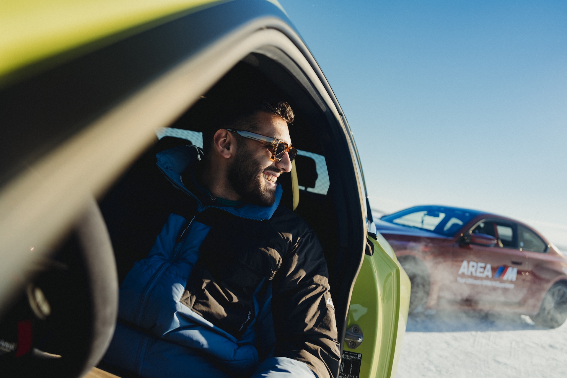 Ein lächelnder Mann mit Sonnenbrille und blauer Jacke sitzt auf dem Fahrersitz eines grünen Fahrzeugs mit offenem Fenster. Ein klarer blauer Himmel ist im Hintergrund sichtbar, und ein weiteres Fahrzeug ist teilweise hinter ihm zu sehen.