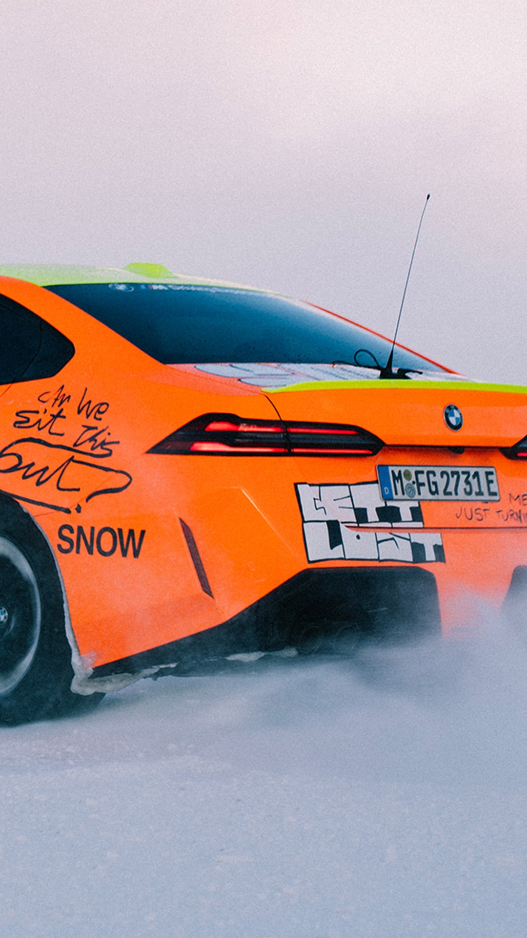 Ein leuchtend orangefarbener BMW Sportwagen, der auf Schnee fährt. Die Rückseite des Fahrzeugs ist sichtbar mit Rücklichtern, einem Kennzeichen und einigen Beschriftungen und Grafiken auf der Karosserie. Das Fahrzeug scheint Schnee aufzuwirbeln, während es sich über eine weiße Schneeoberfläche bewegt.