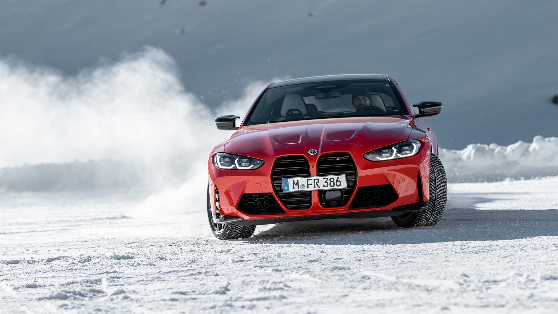 Ein leuchtend roter BMW Sportwagen mit dem Kennzeichen M-FR 386 fährt auf einer schneebedeckten Oberfläche. Der markante Kühlergrill und die Scheinwerfer des Fahrzeugs sind deutlich sichtbar, während es durch das verschneite Gelände mit weißen Schneeverwehungen im Hintergrund fährt.
